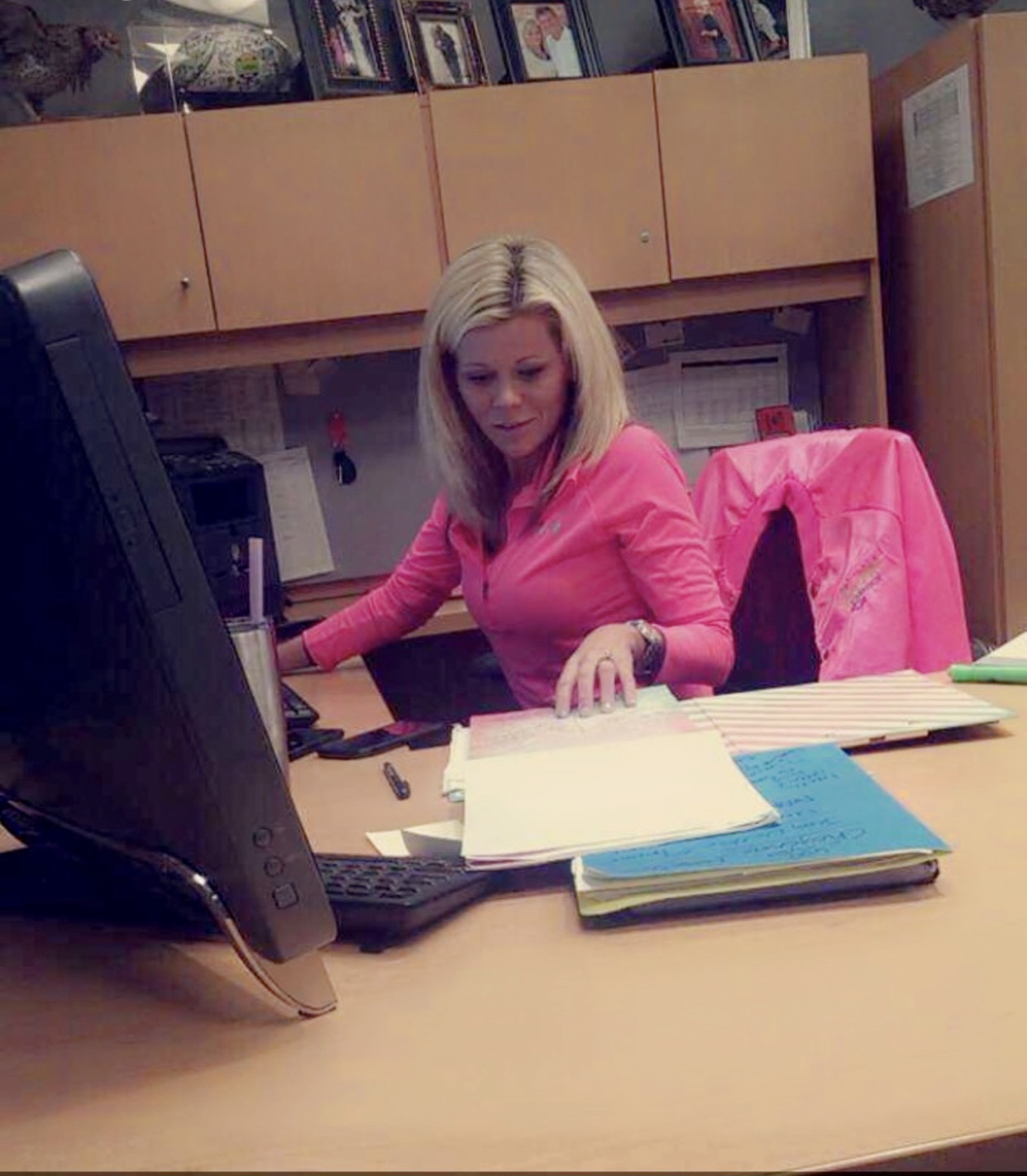 Woman in pink shirt sitting at desk with papers, computer, and pink jacket behind her, in an office setting.