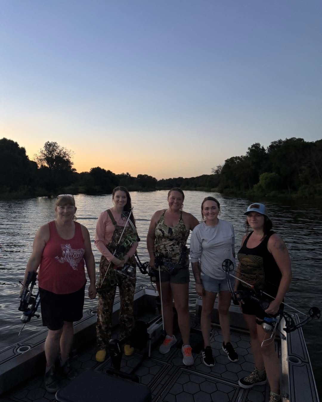 Five women stand on a boat by a river at sunset, smiling and posing for the photo.