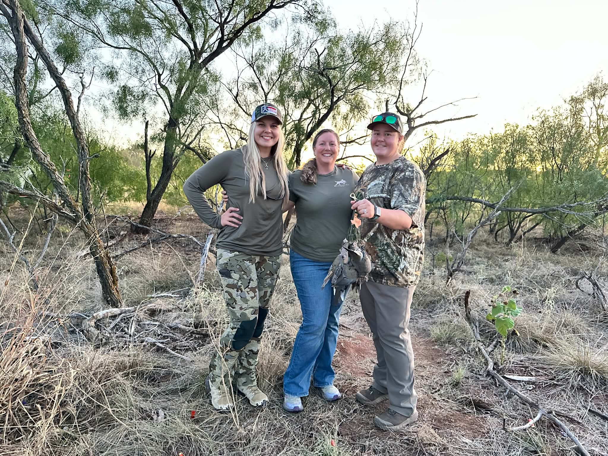 Three people standing outdoors among trees and dry grass, smiling and posing for the photo.