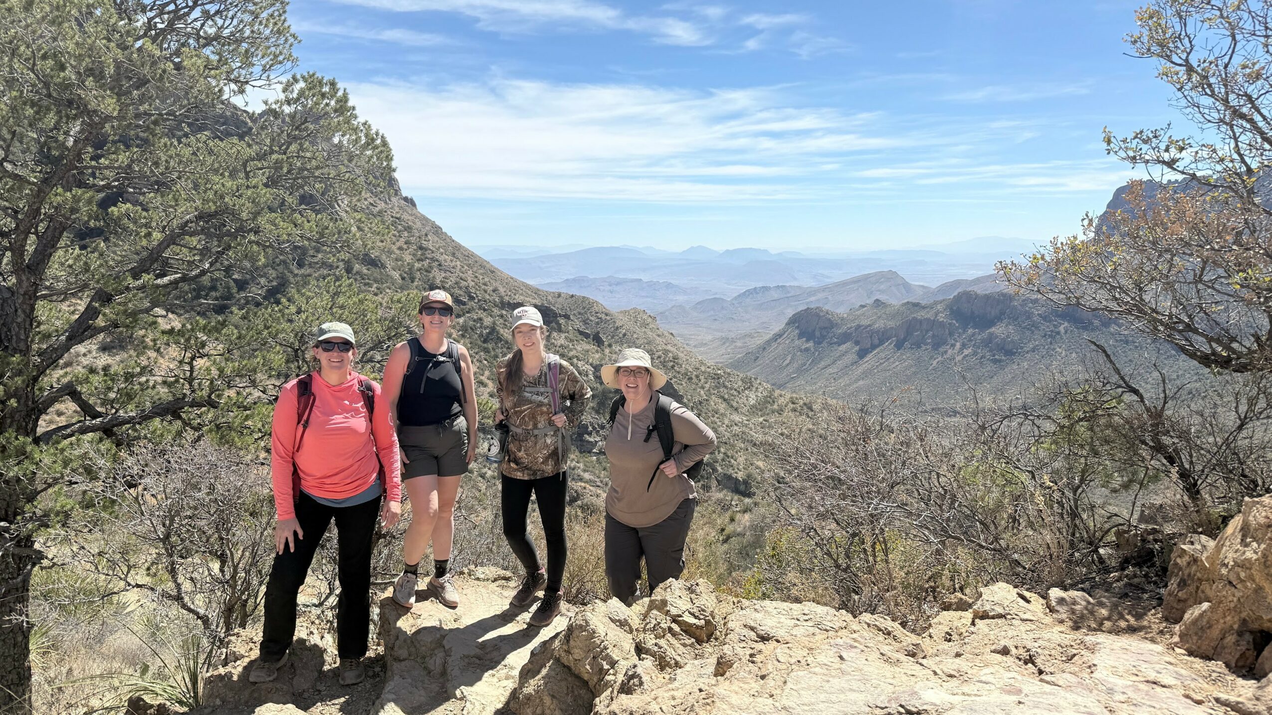 Four people hiking on a trail with mountains and trees in the background.