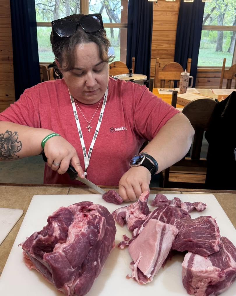 Woman cutting raw meat on a white cutting board in a room with wooden walls and large windows, wearing a red shirt and glasses on head.