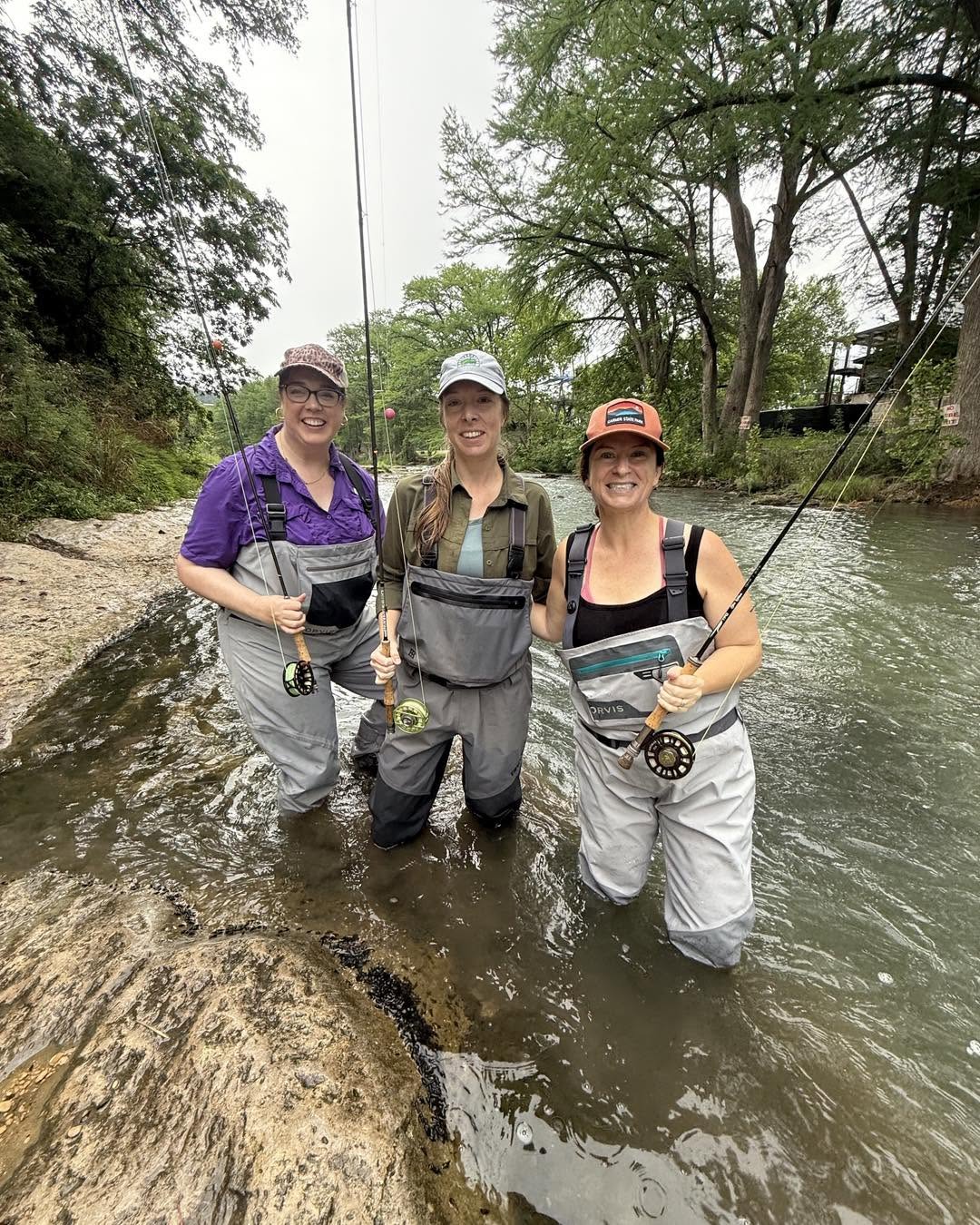 Three women stand in a river holding fishing rods, smiling, with trees and a house in the background.