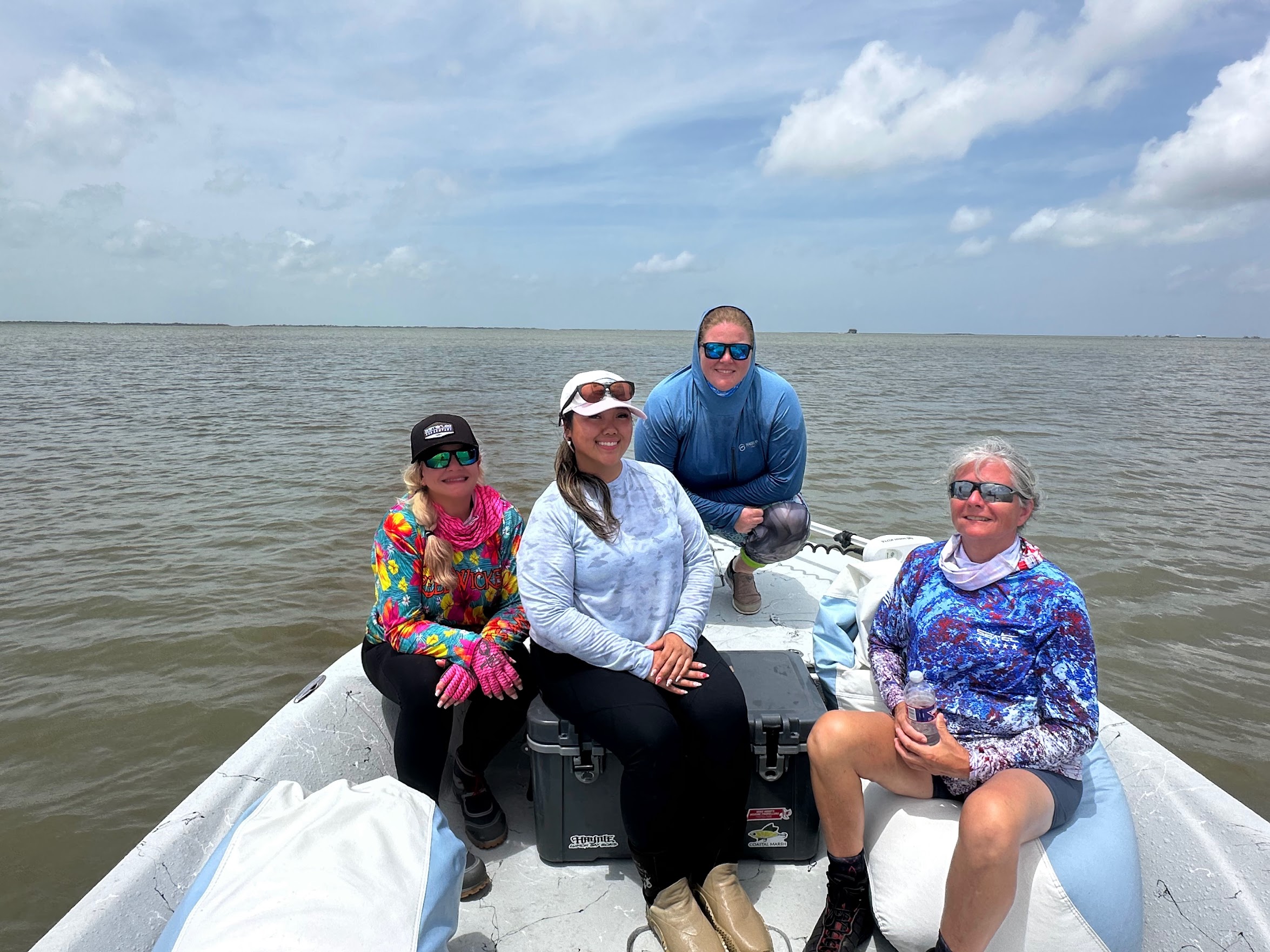 Four people sitting on a boat in water, smiling, wearing sunglasses and colorful clothing, with a cloudy sky background.