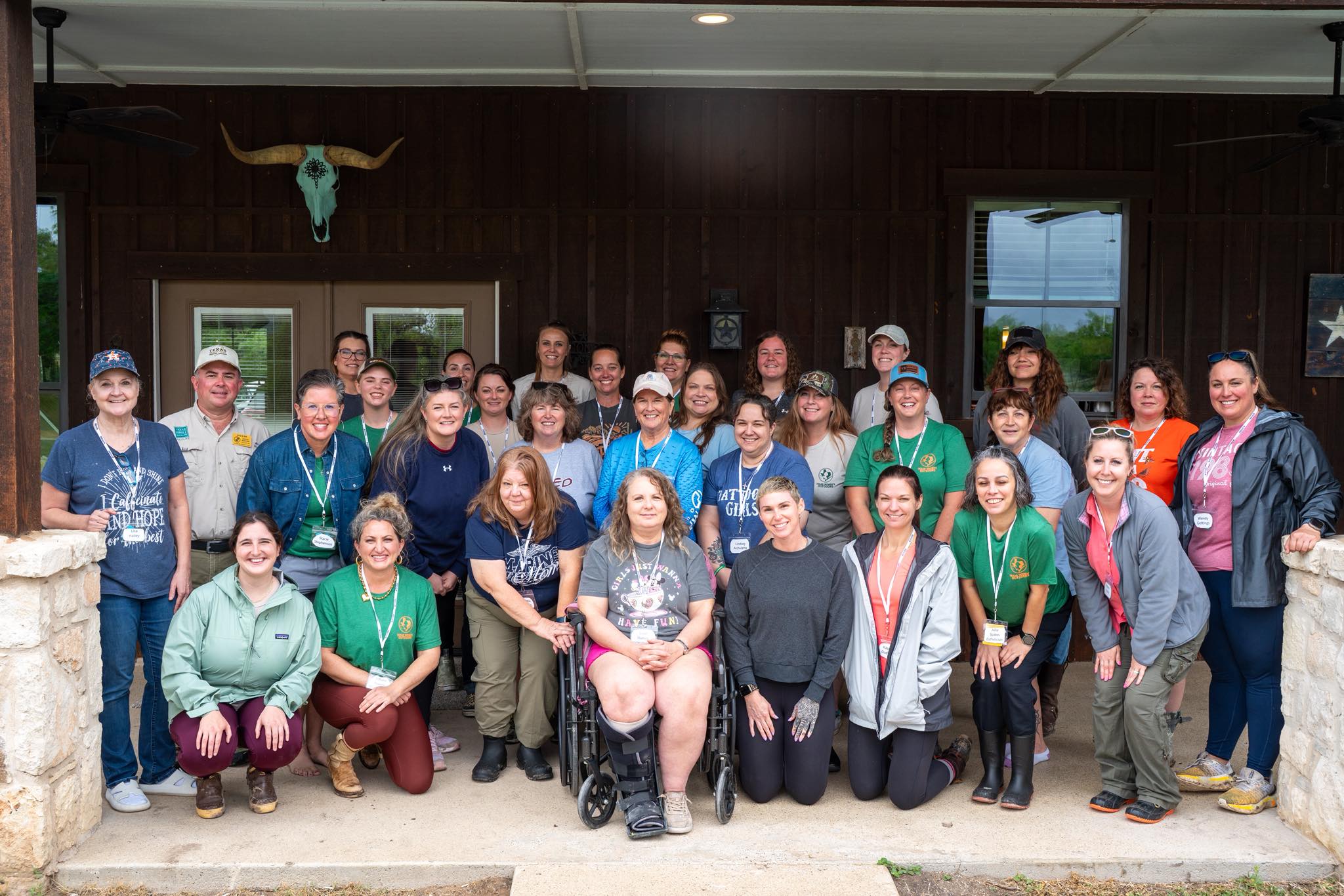 Group of people posing outdoors in front of a dark wooden building, some wearing green shirts, smiling.