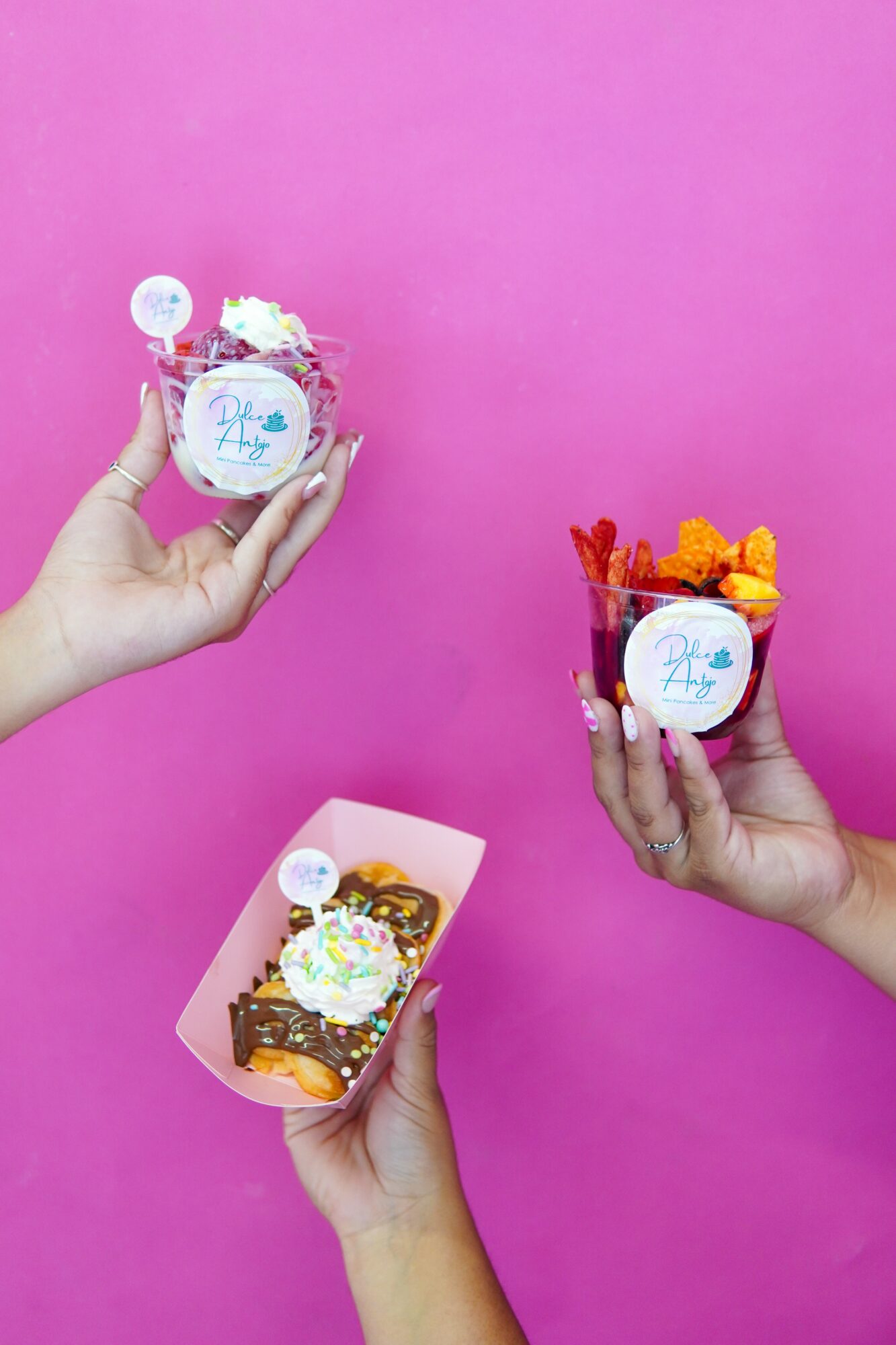 Three hands holding cups and a tray with colorful desserts against a pink background.