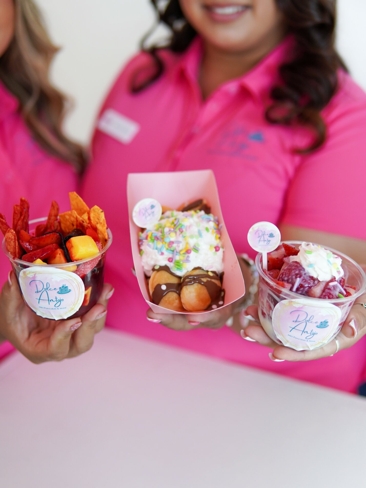 Two people in pink shirts hold cups with colorful desserts and toppings, smiling, against a pink background.