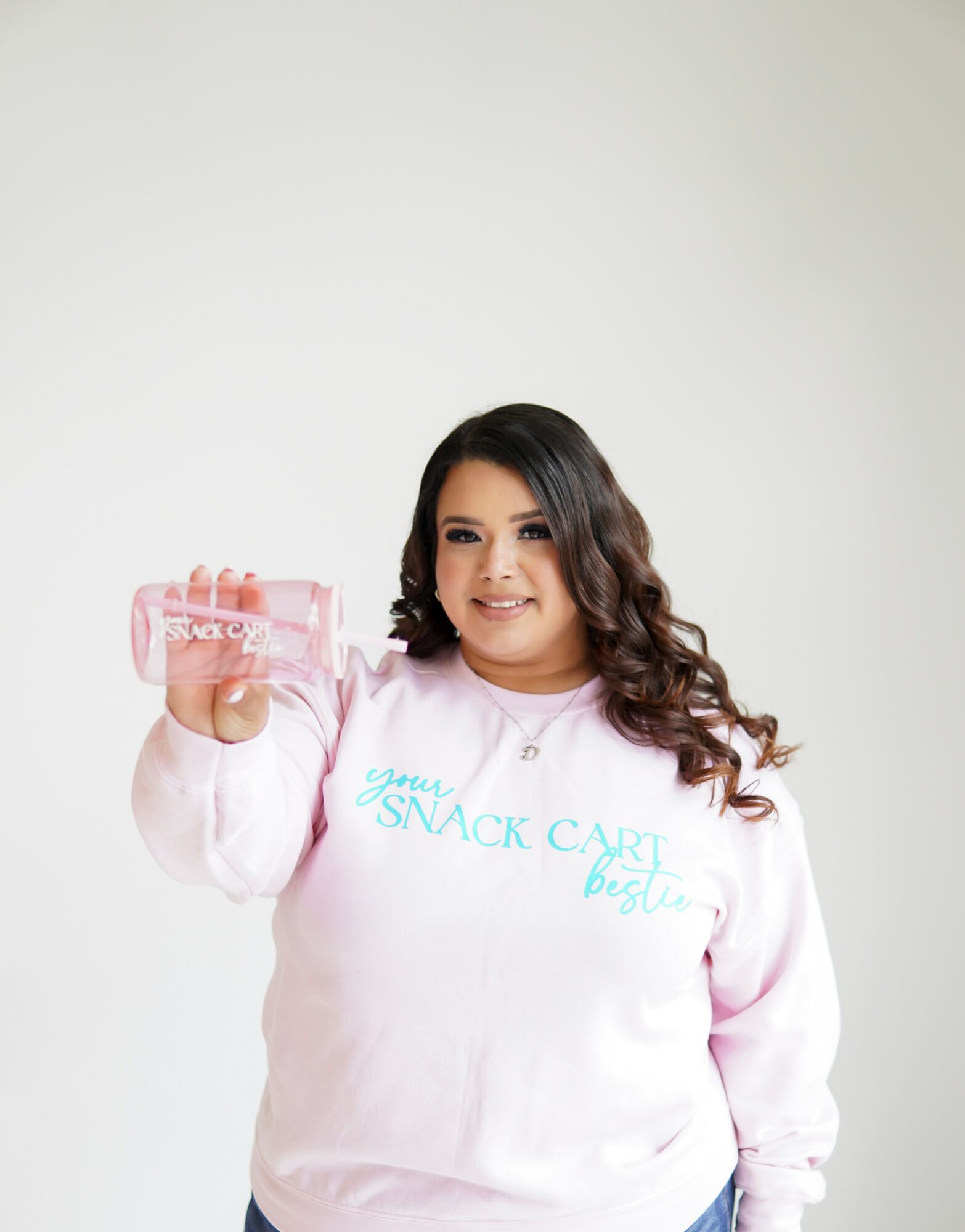 Woman with long dark hair holding a pink snack container, smiling, wearing a light pink sweatshirt with blue text, standing against a plain background.