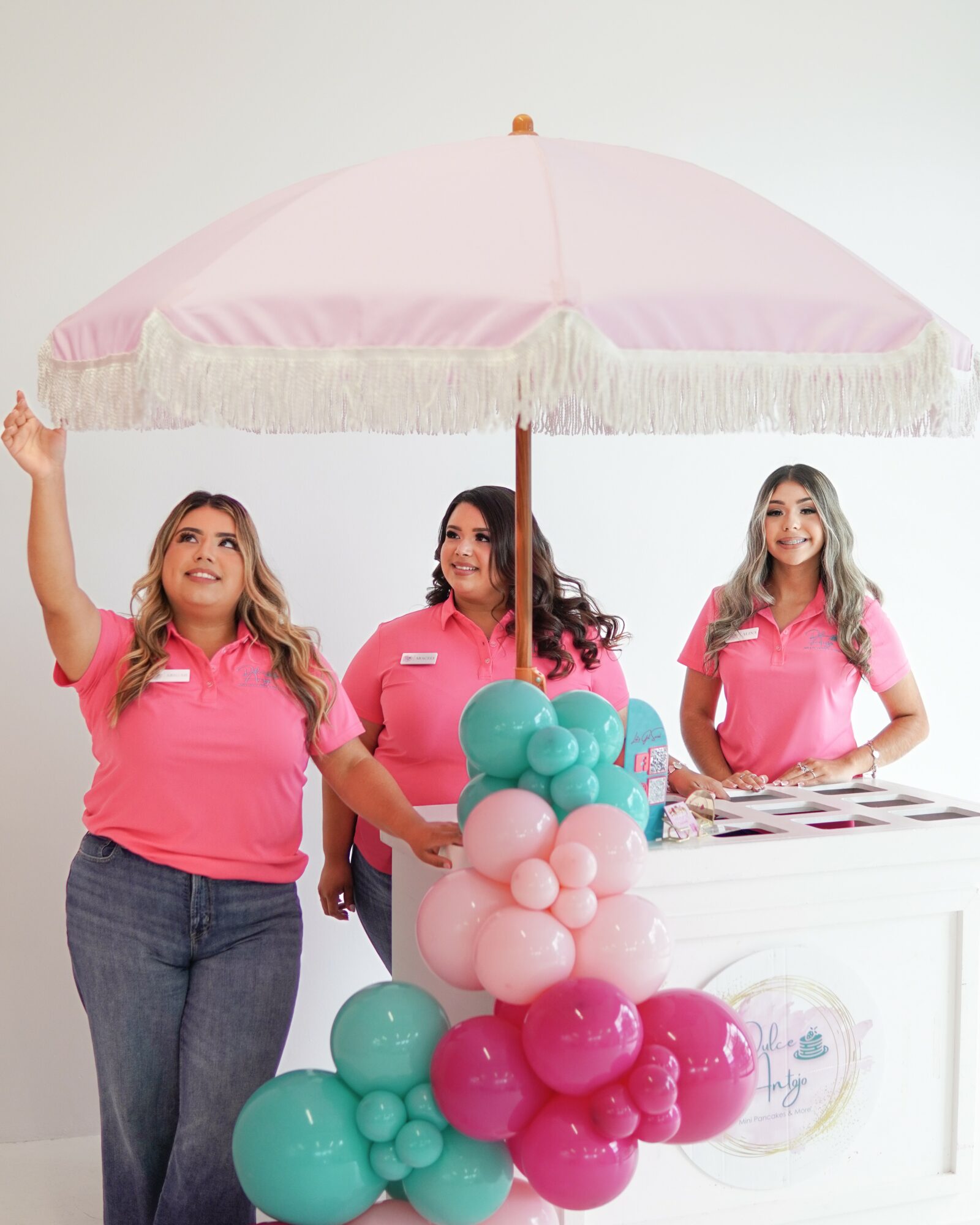 Three women in pink shirts stand behind a white counter with a pink umbrella decorated with balloons, smiling and waving.