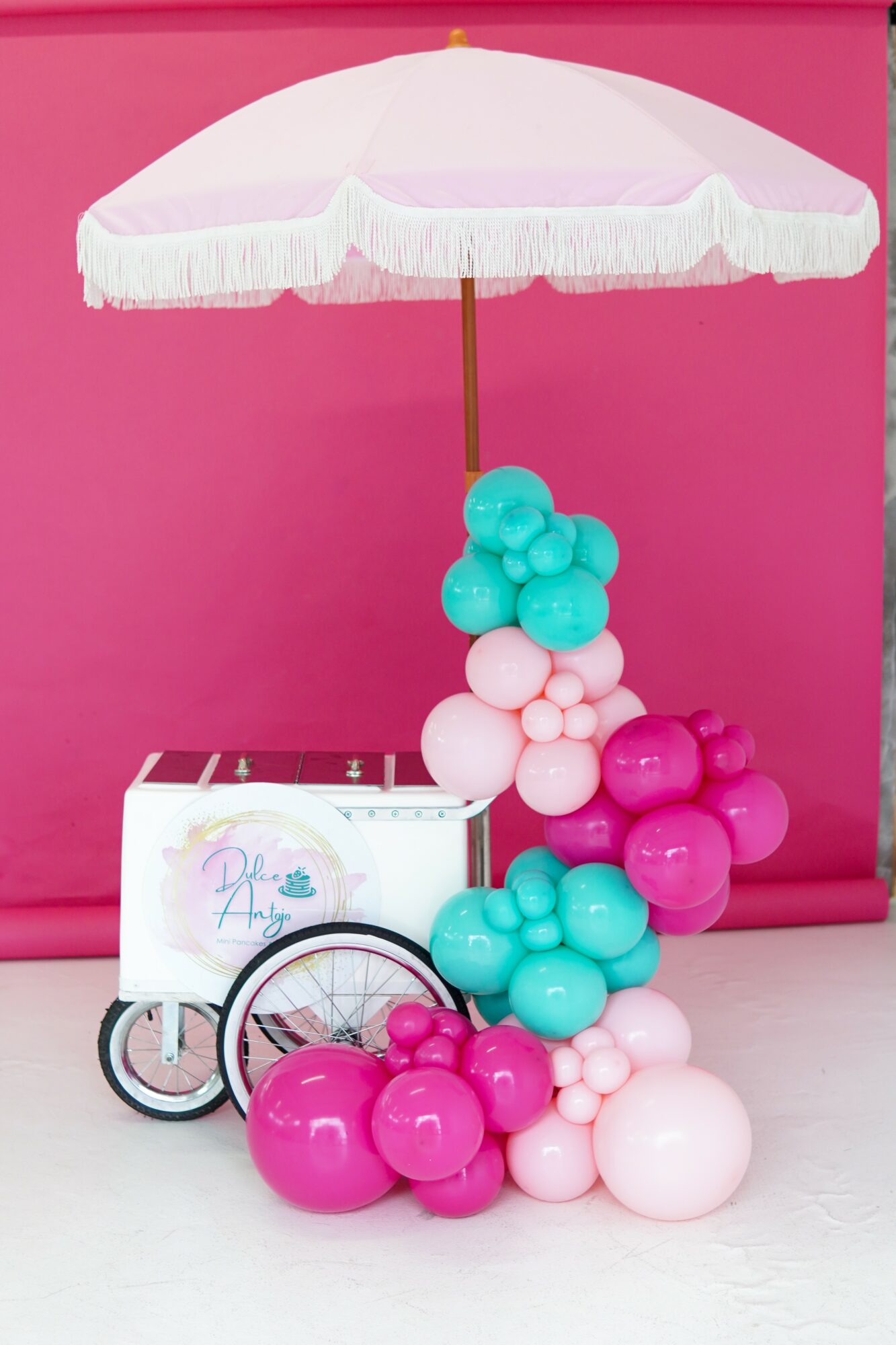 Pink and white balloon arrangement with a pink and white umbrella and a small cart against a pink background.