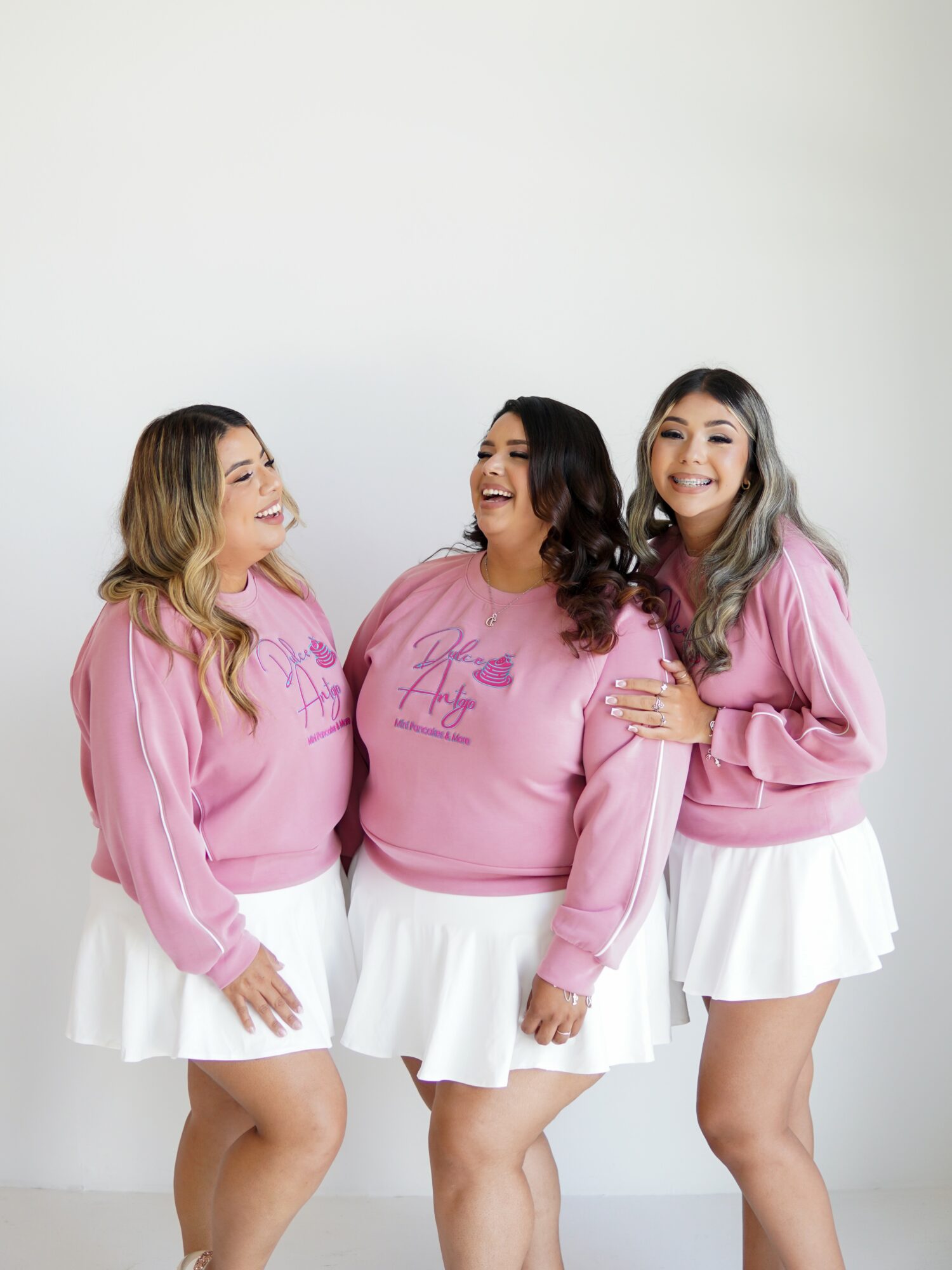 Three women in pink sweatshirts and white skirts smiling and talking together against a plain white background.