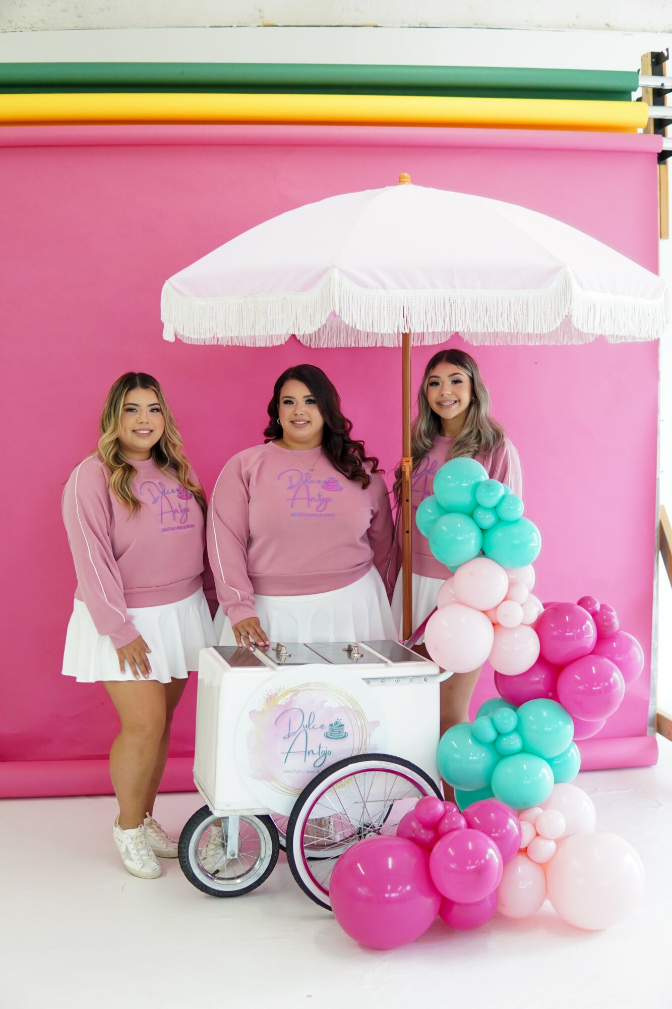 Three women in pink tops and white skirts stand beside a pink and white balloon display with a white umbrella, pink backdrop, and colorful overhead stripes.