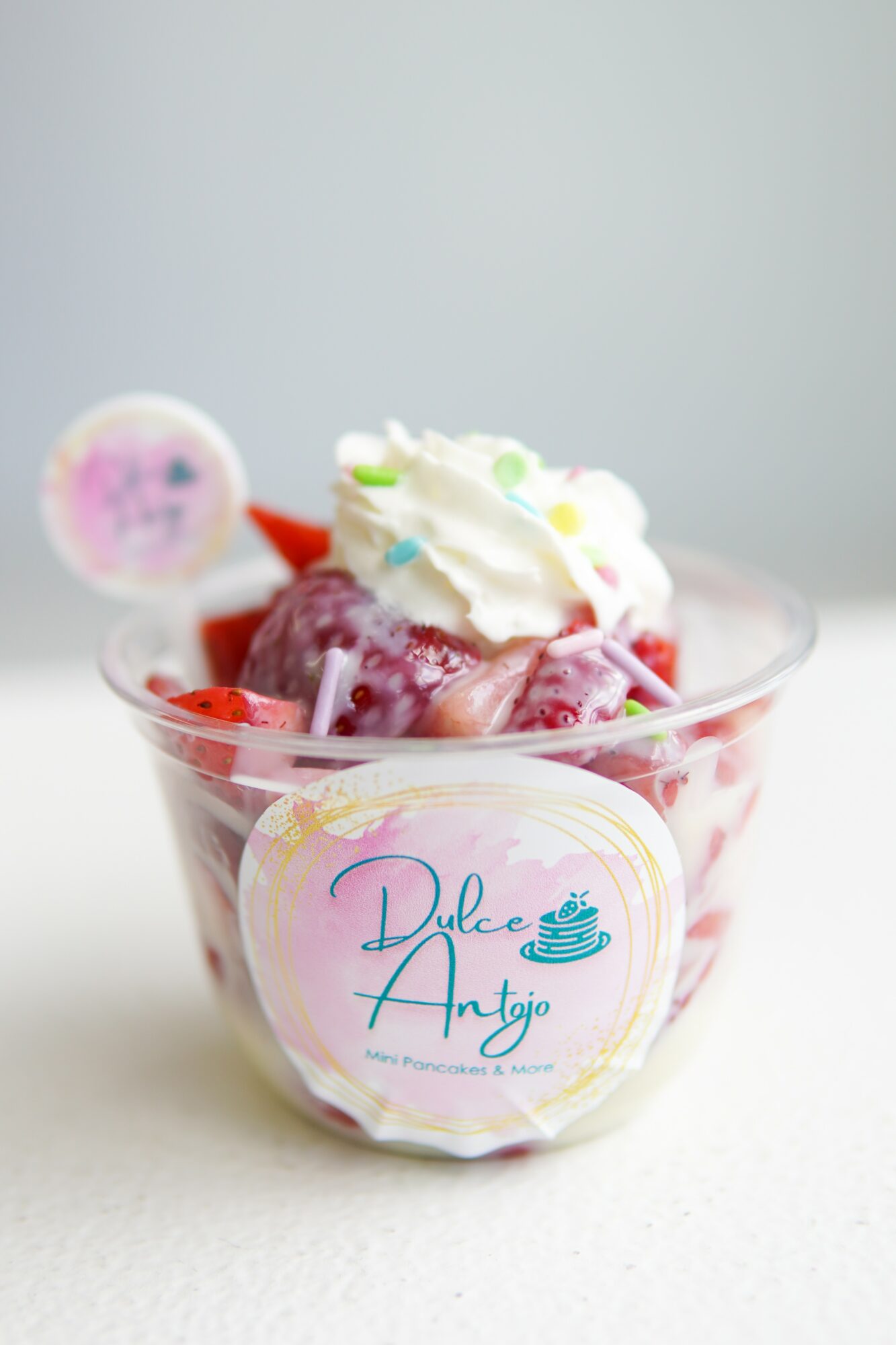 Cup of strawberries topped with whipped cream and green sprinkles, with a pink and white spoon in the background.