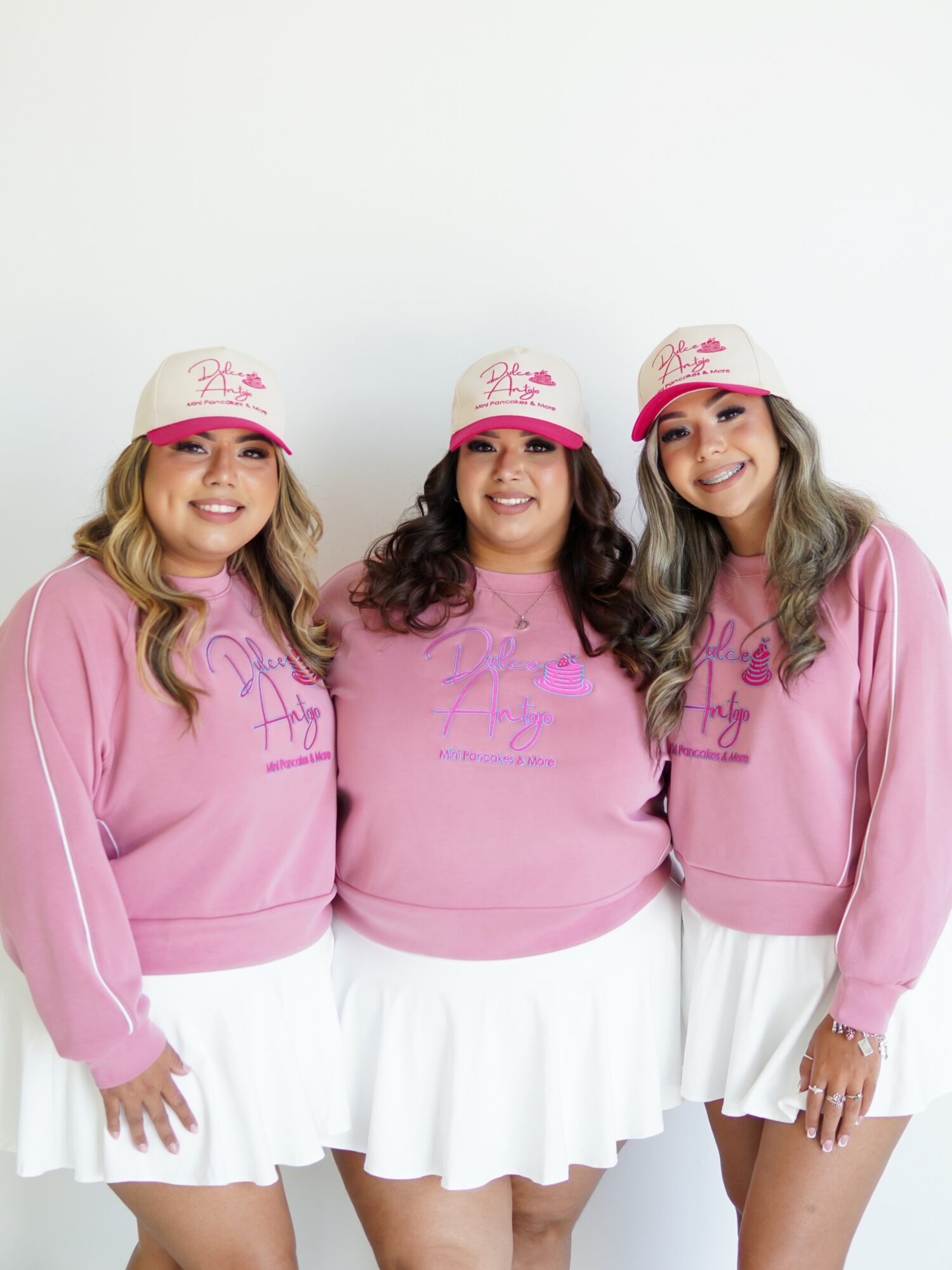 Three women in pink sweatshirts and white skirts, wearing pink hats, smiling and standing together against a white background.