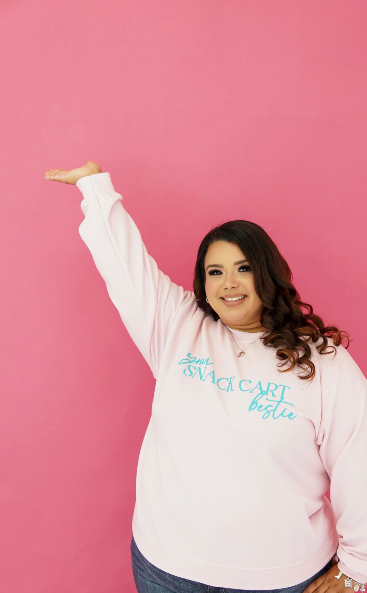 Smiling woman with long dark hair in a white sweatshirt against pink background, raising her right arm.