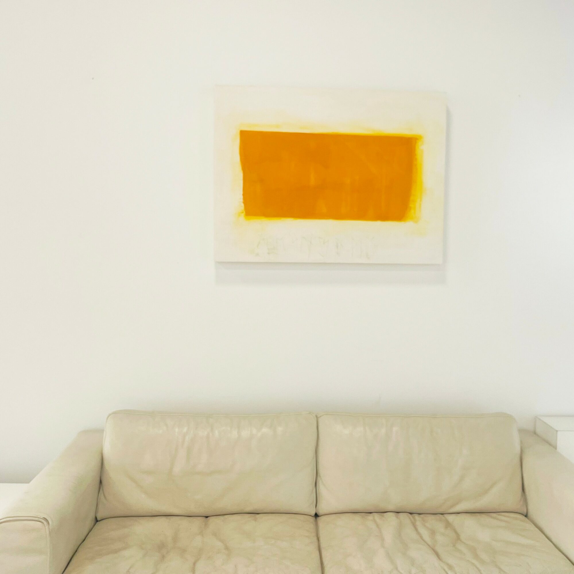 White wall with a rectangular yellow-orange abstract painting above a beige sofa, minimalistic interior design.