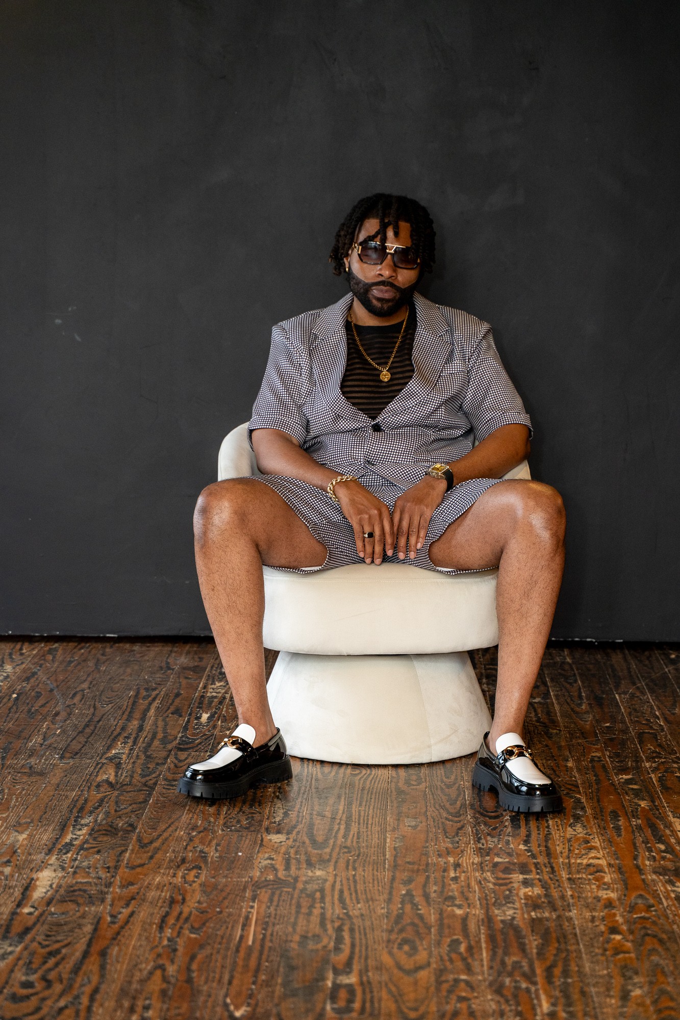 Person sitting on a white chair with legs apart, wearing sunglasses, a patterned blazer, shorts, and black shoes, against a dark wall.