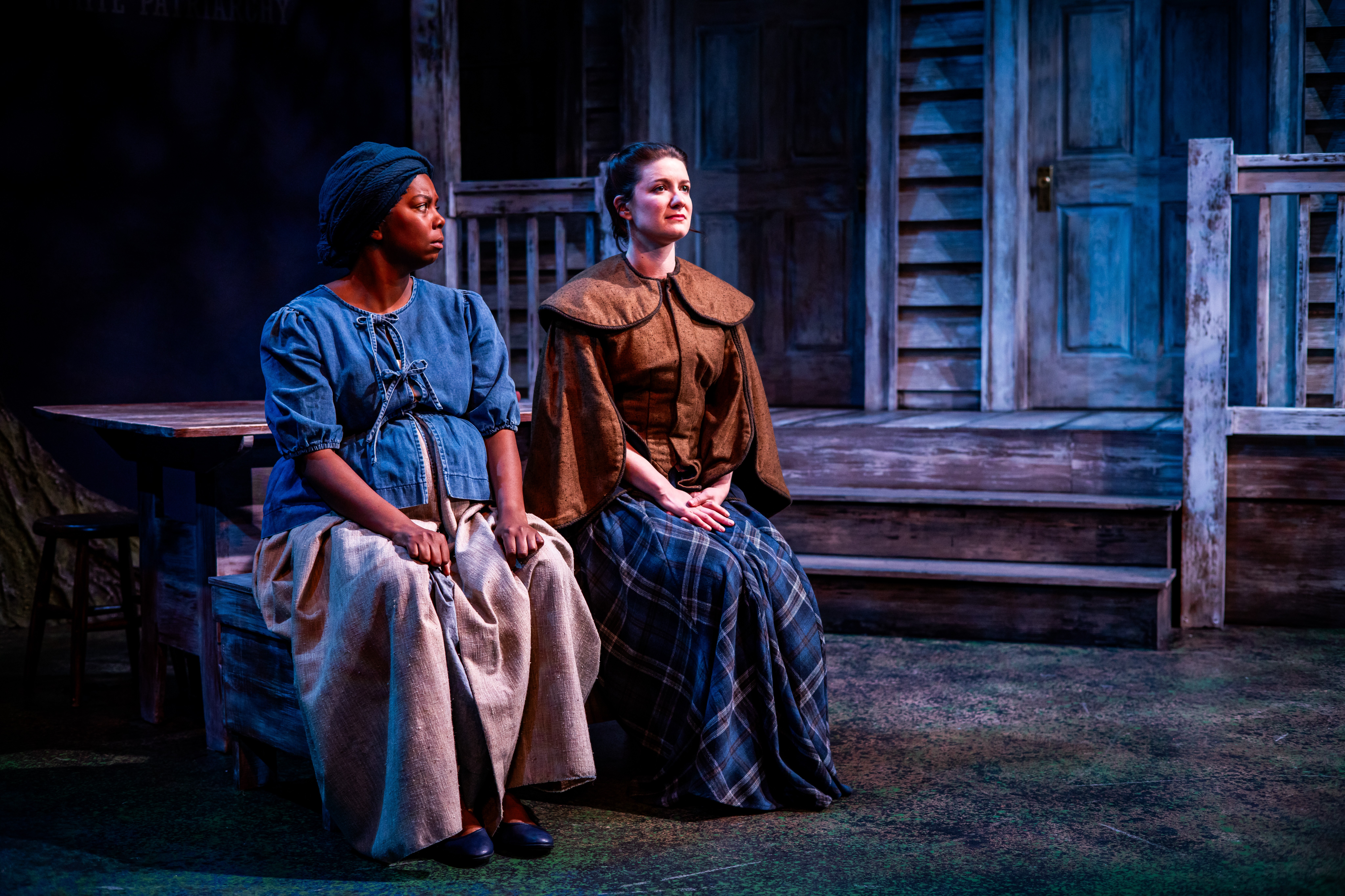 Two women sit on a bench outdoors in front of a wooden building, dressed in period clothing, with a somber expression.