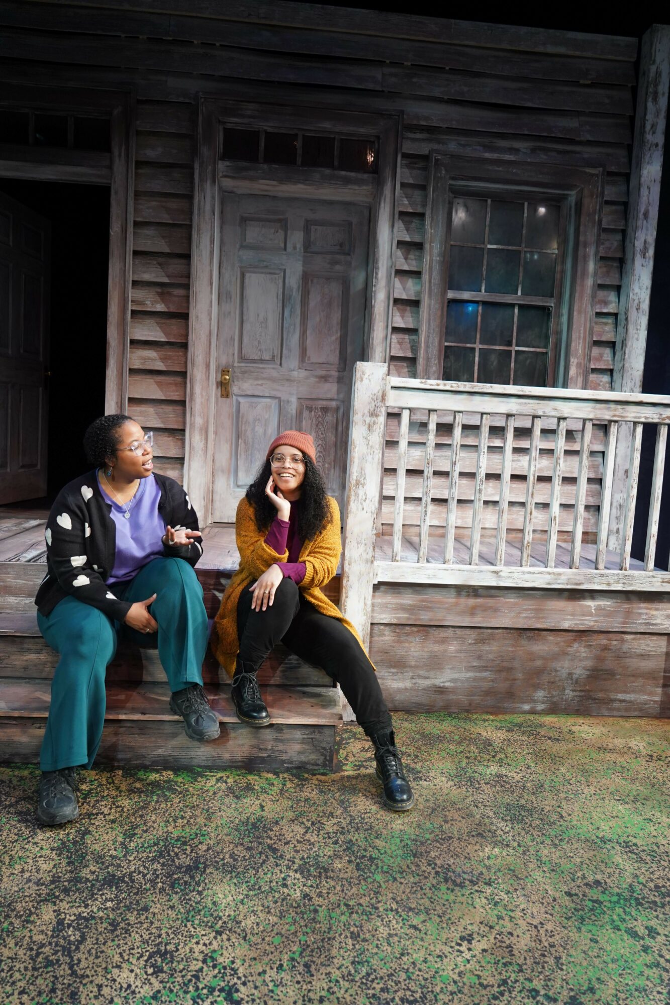 Two women sitting on a wooden porch, one in a purple shirt and the other in a yellow shirt, with a wooden door and railing behind them.