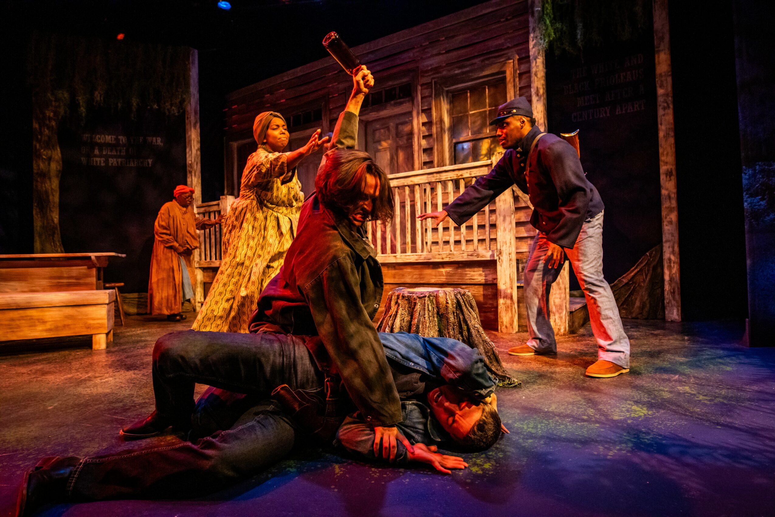 Theater scene with actors in period costumes on stage, some standing and others on the floor, with a wooden house backdrop.