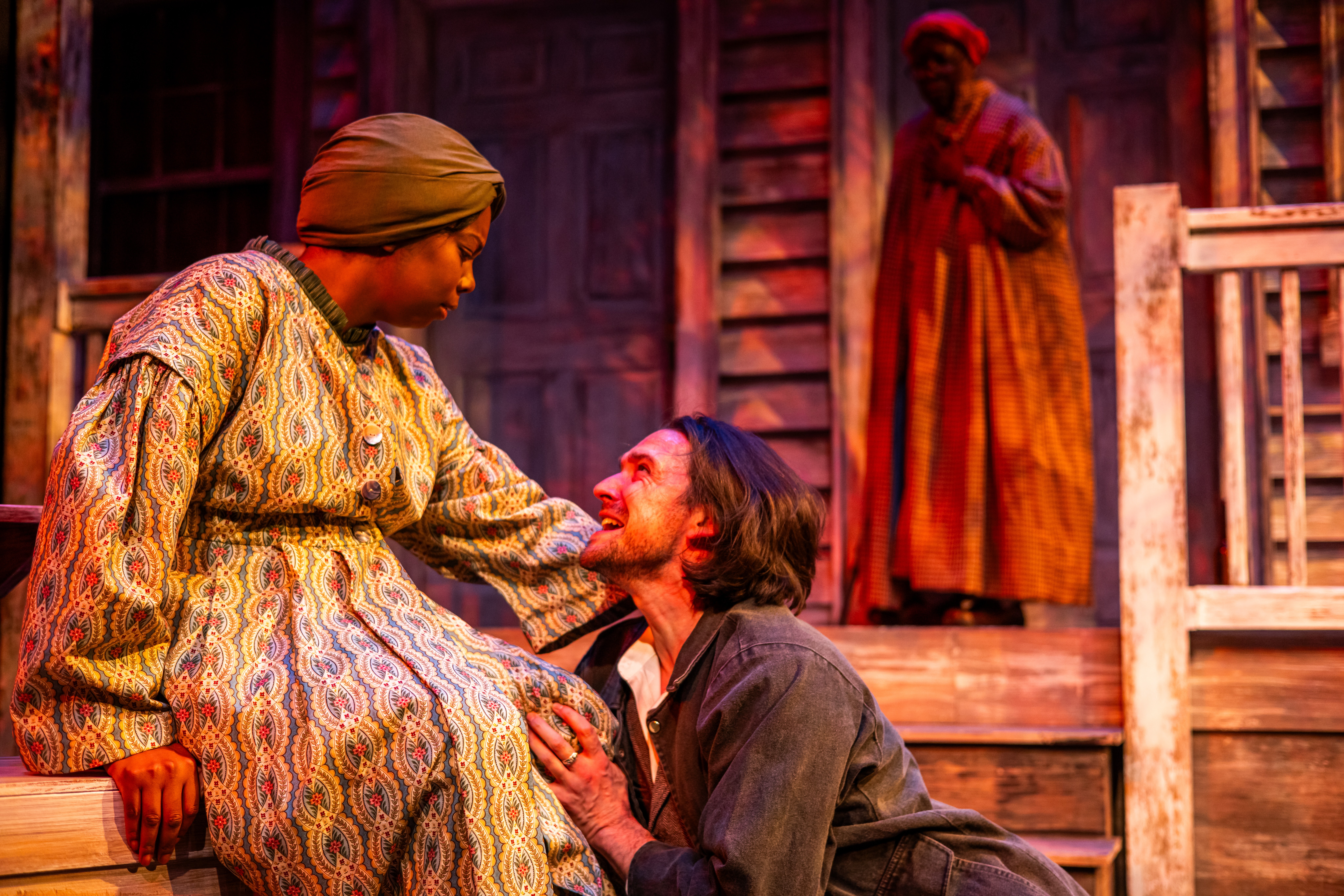 Two women on stage, one kneeling and touching the other's face, with a third woman in the background. Stage set resembles a rustic house.
