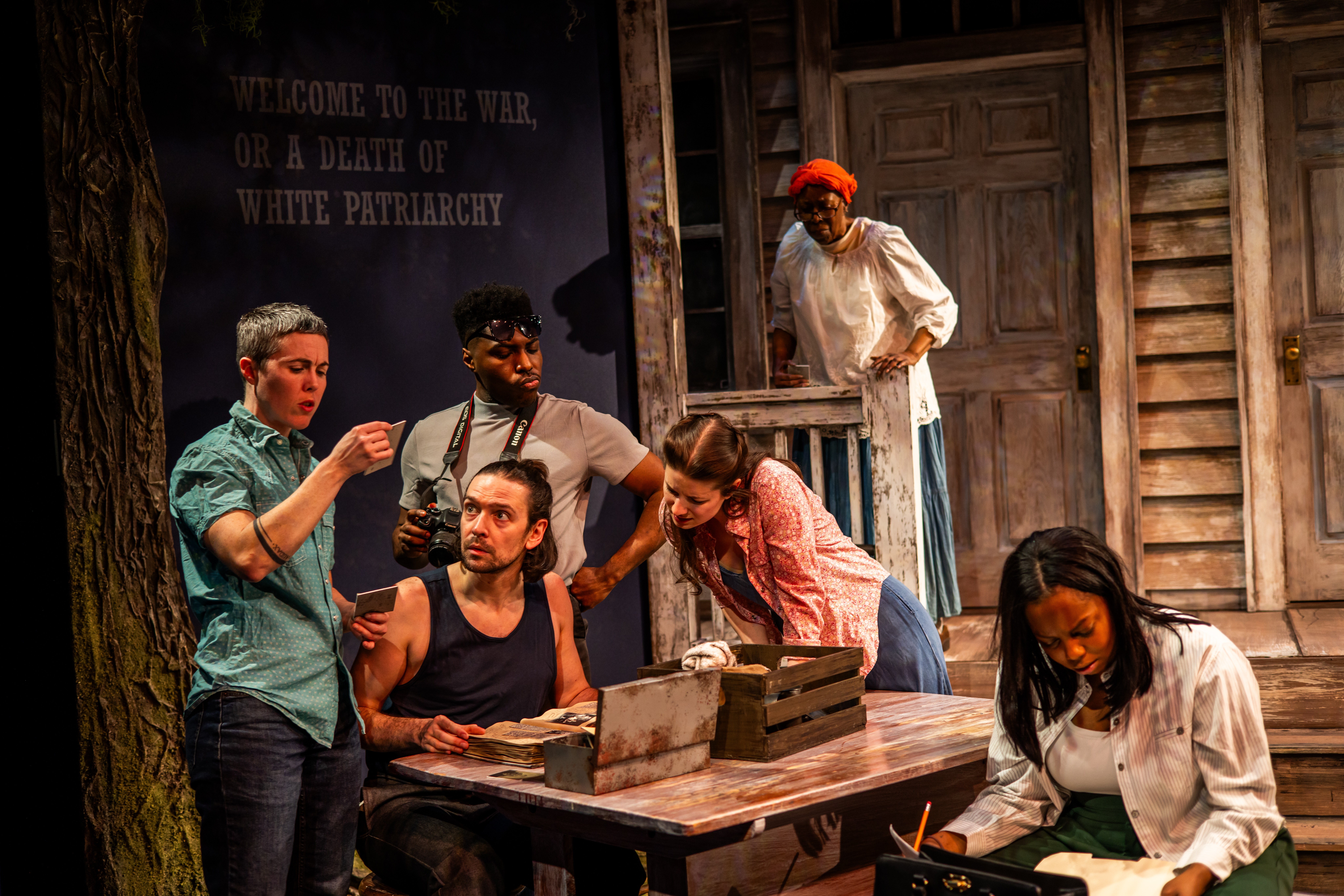 Group of six people around a table on stage, with a wooden backdrop and a sign about patriarchy, engaged in discussion.