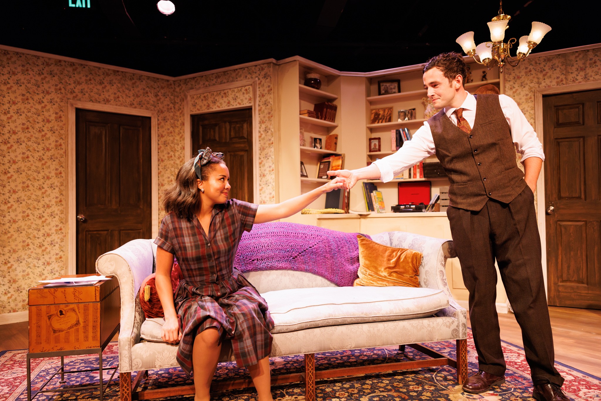 Man and woman on stage, woman sitting on sofa, man reaching out hand, stage set with furniture and books, warm lighting.
