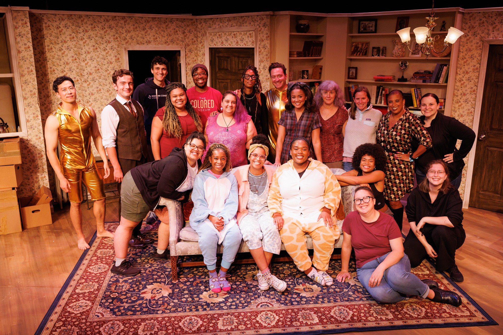 Group of 17 people gathered in a living room, some sitting on a sofa and others standing behind, smiling at the camera.