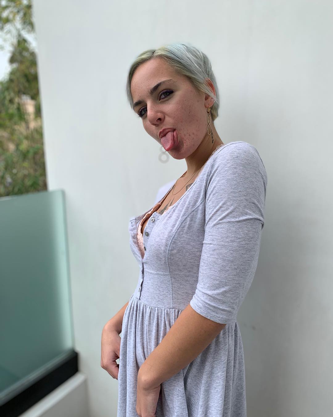Young woman with blonde hair sticking out her tongue, wearing a light gray dress, standing against a plain wall.