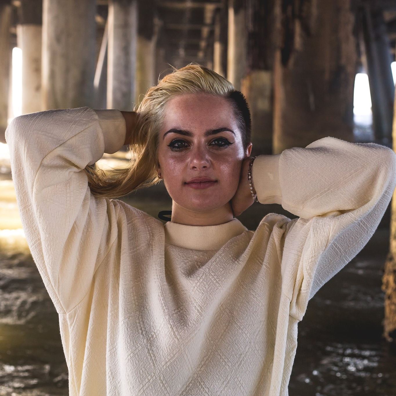 Woman with blonde hair and makeup, wearing a cream sweater, standing under a pier with wooden supports and water below.
