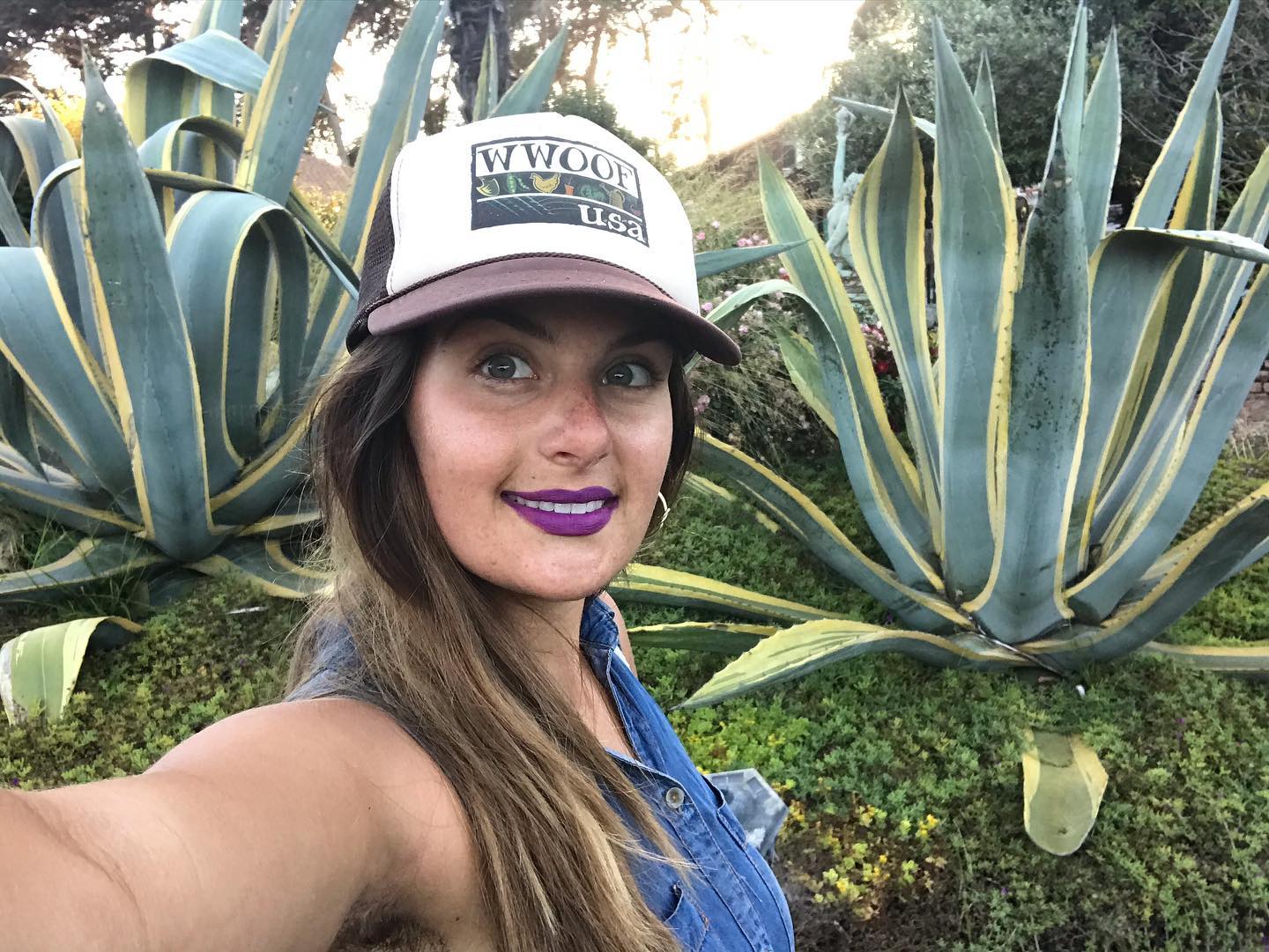 Woman taking selfie outdoors with large agave plants in background, wearing a cap and denim shirt, smiling.