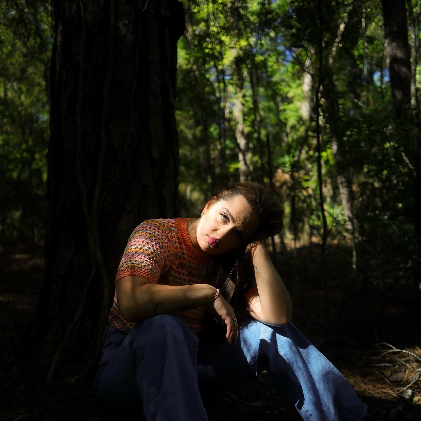 Young woman sitting in a forest, leaning against a tree, with sunlight filtering through leaves.
