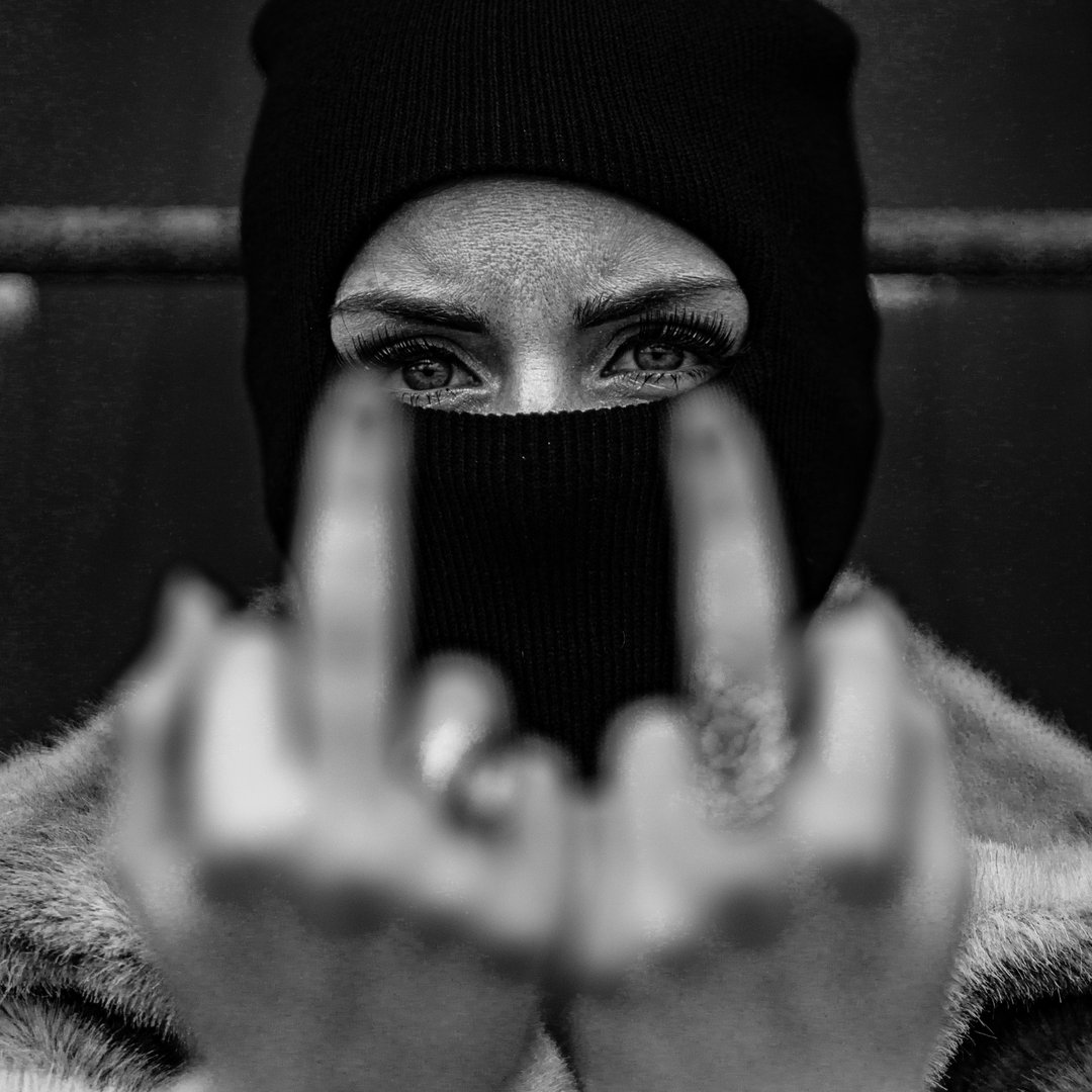 Person wearing a black beanie and face covering, eyes visible, holding up middle fingers towards the camera.