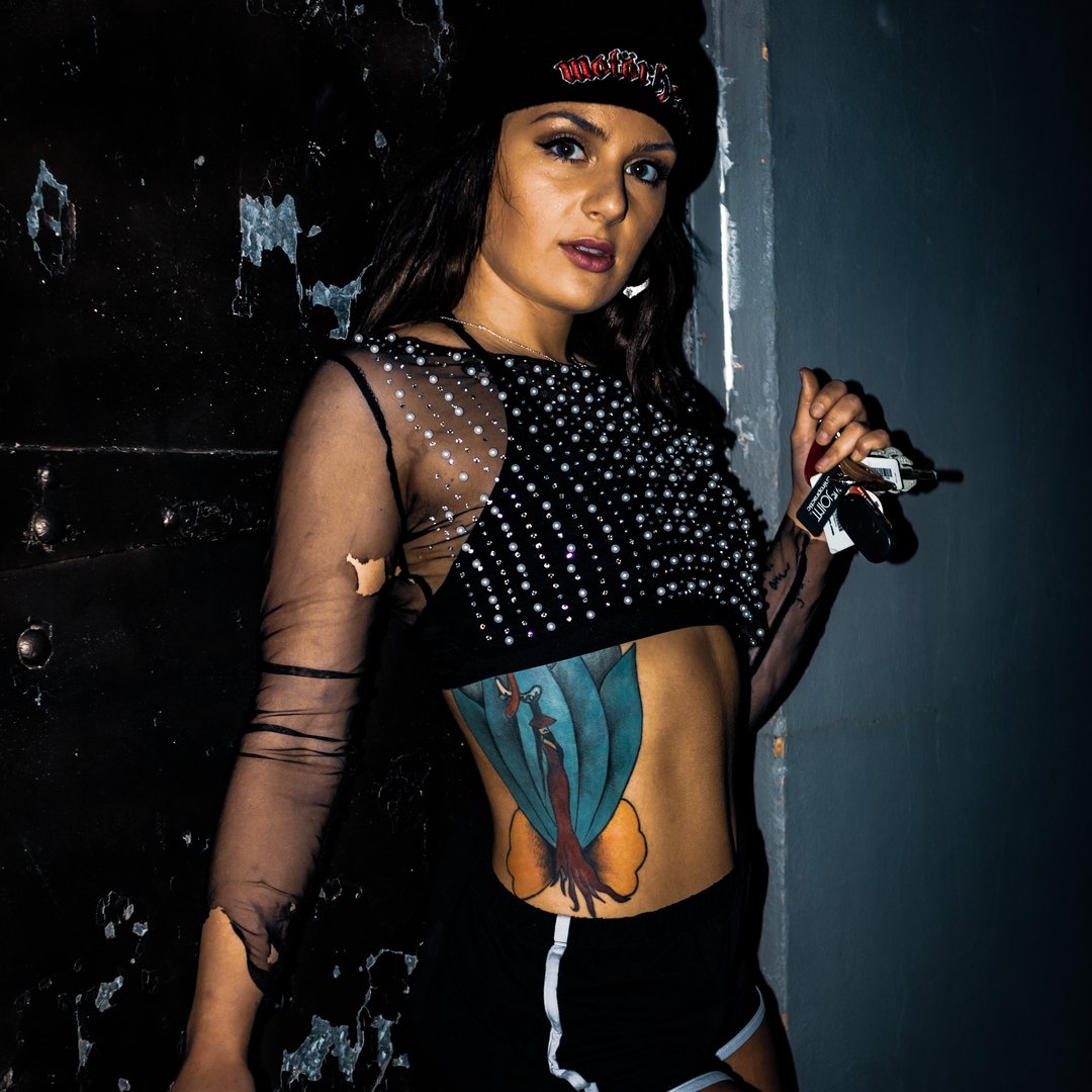 Woman with dark hair, wearing a beanie, black mesh top, and shorts, holding a camera, standing against a dark wall.
