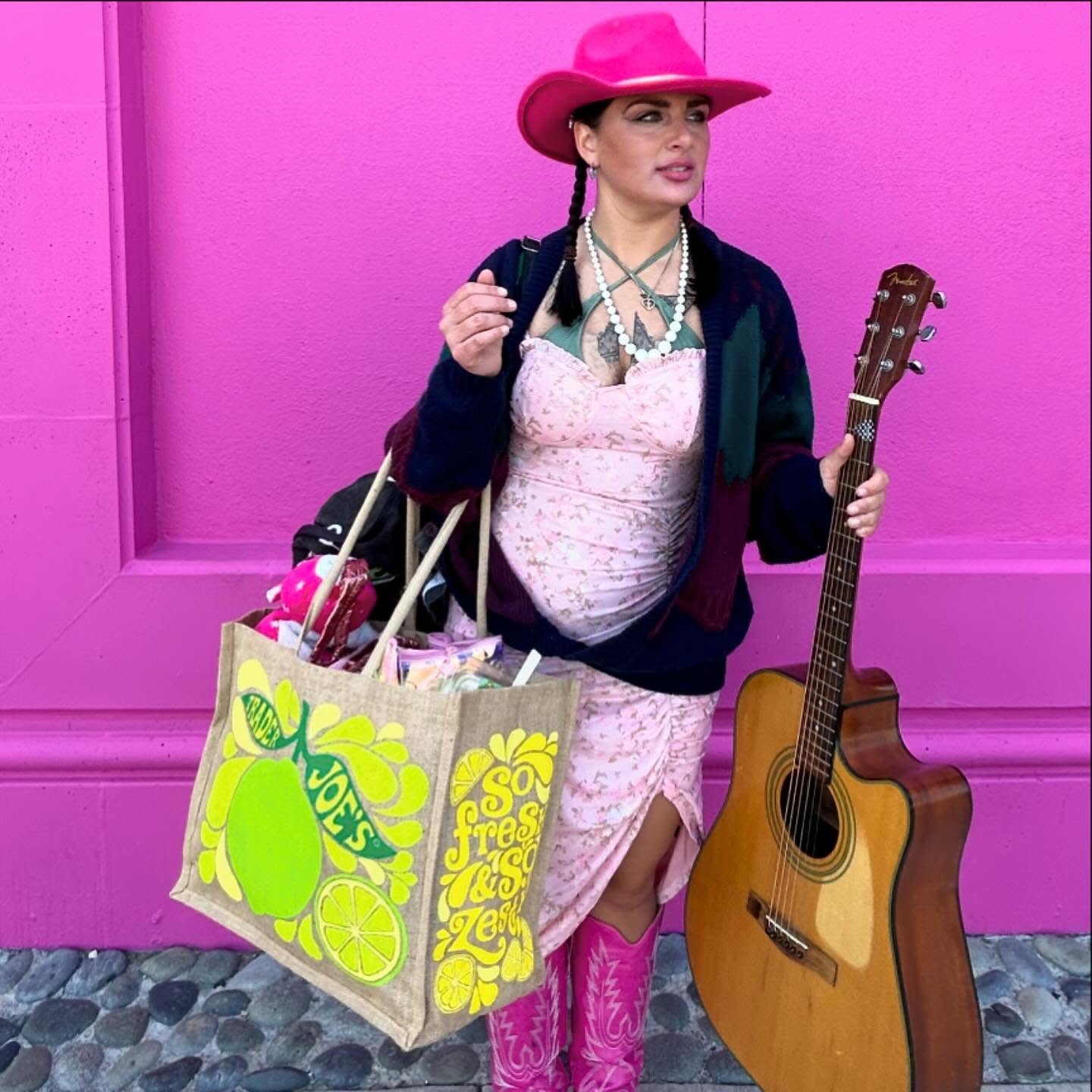 Woman in pink hat and dress holding a guitar and shopping bags against a pink wall.