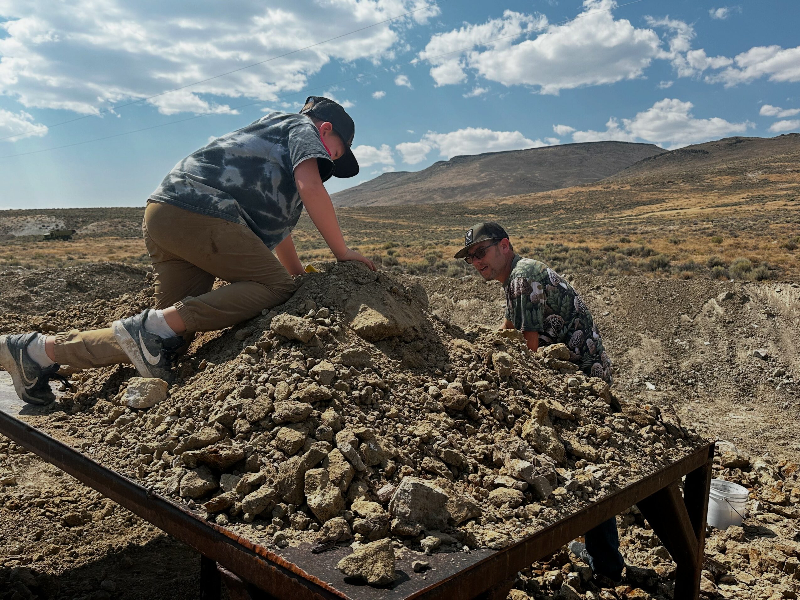 Two people digging in a rocky outdoor landscape with hills and a partly cloudy sky.