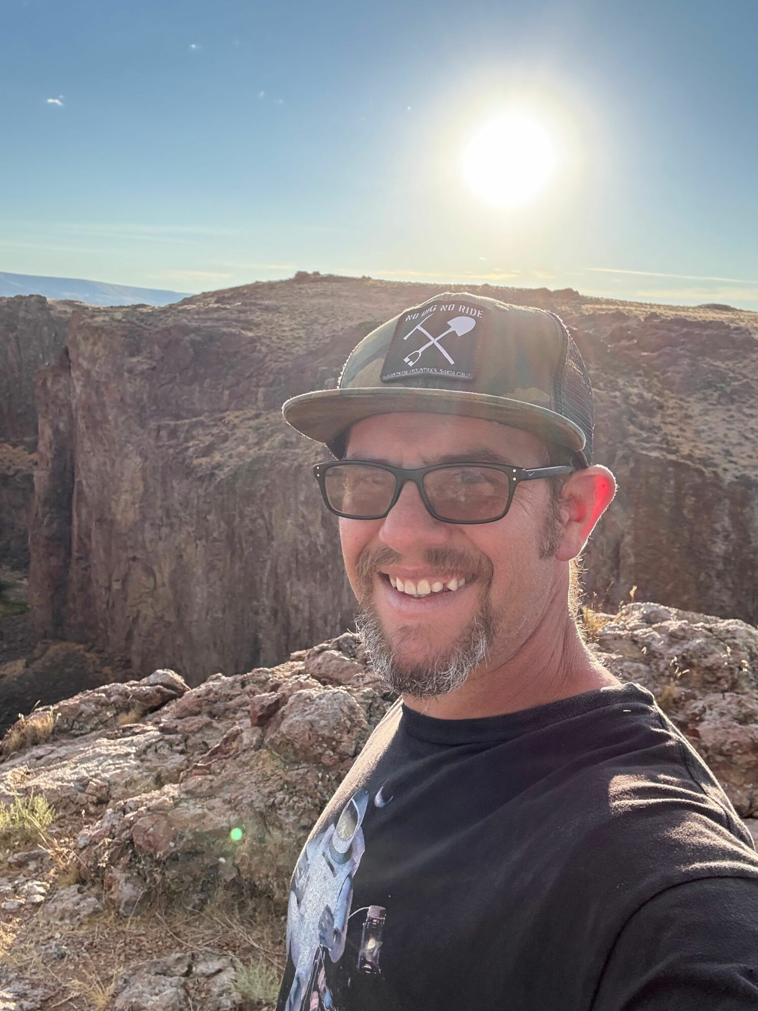 Man with glasses and a cap smiling outdoors with rocky cliffs and a bright sun in the background.