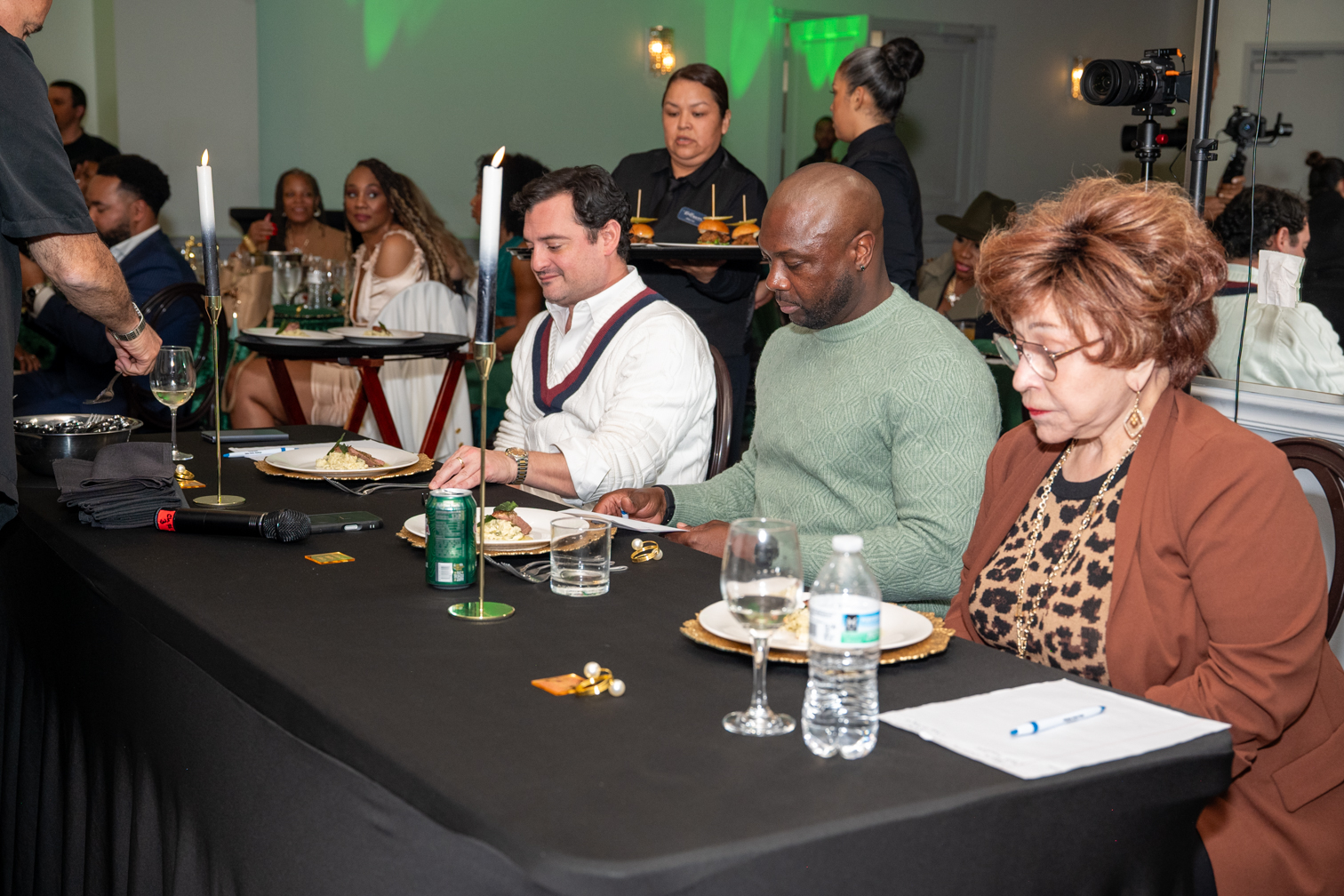 Three people seated at a table with drinks and papers, in a busy indoor event setting.