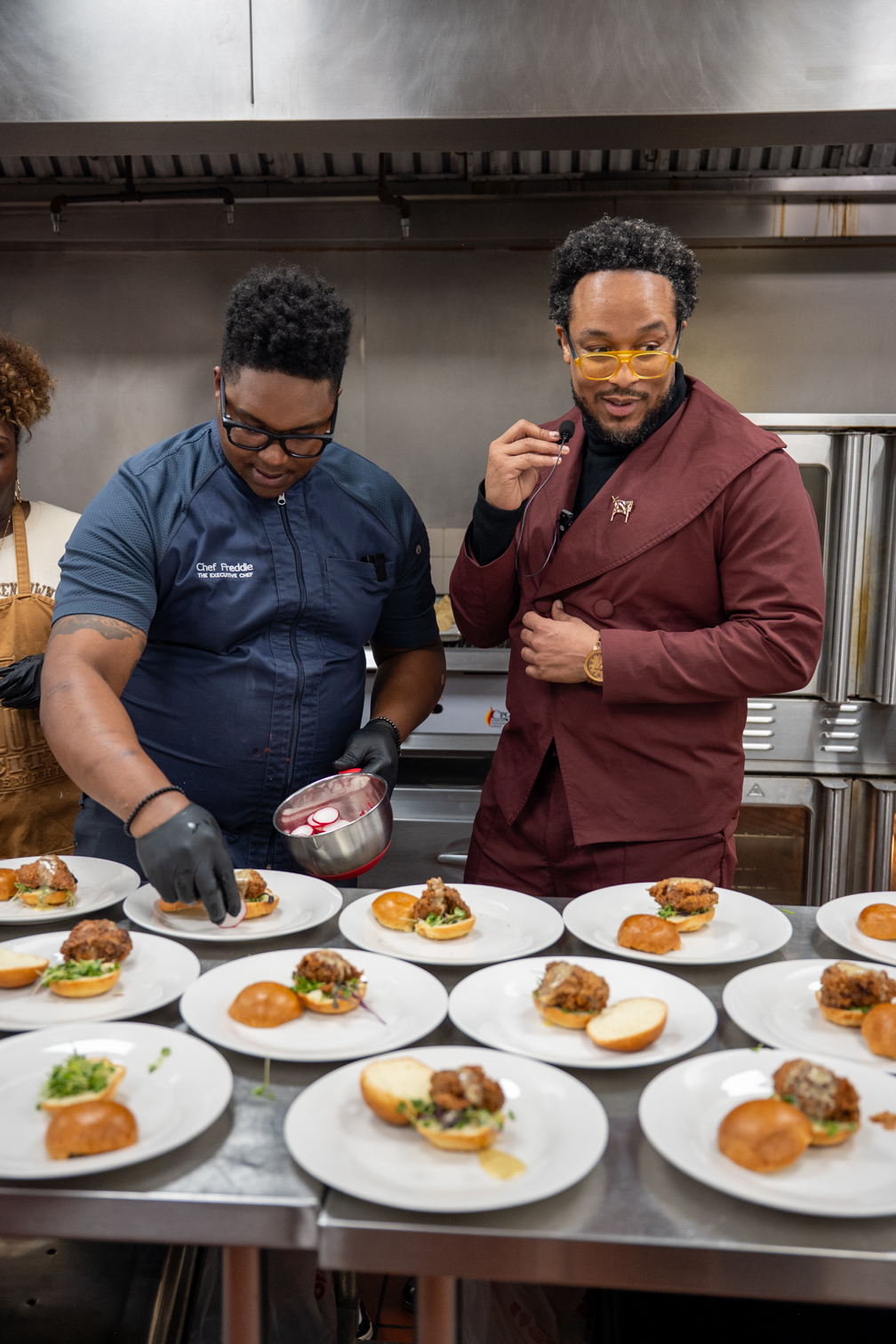 Two chefs prepare plated dishes in a professional kitchen, with multiple plates of food on the counter.