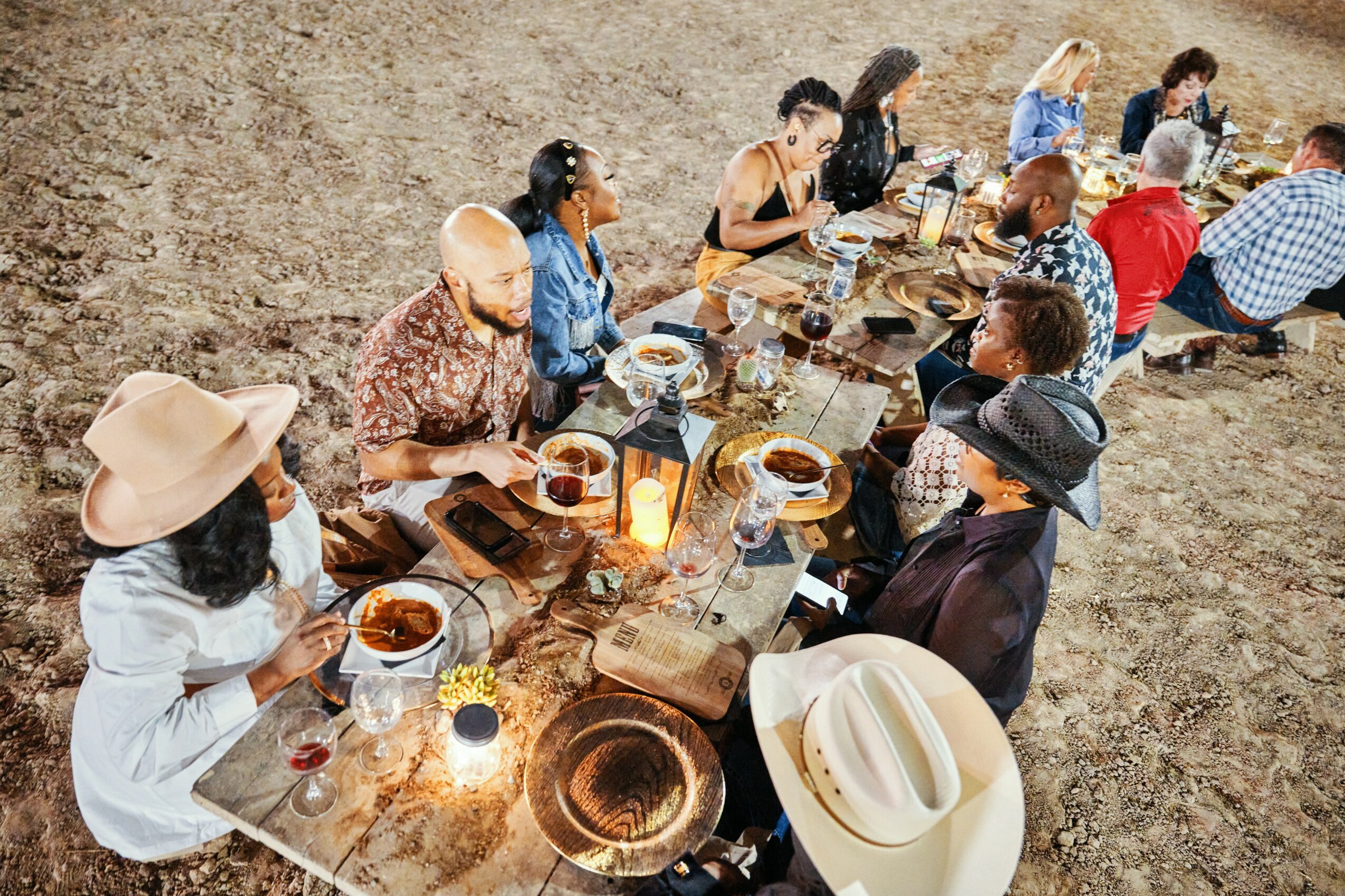 People sitting at a long outdoor table eating food, some wearing hats, on a dirt surface.