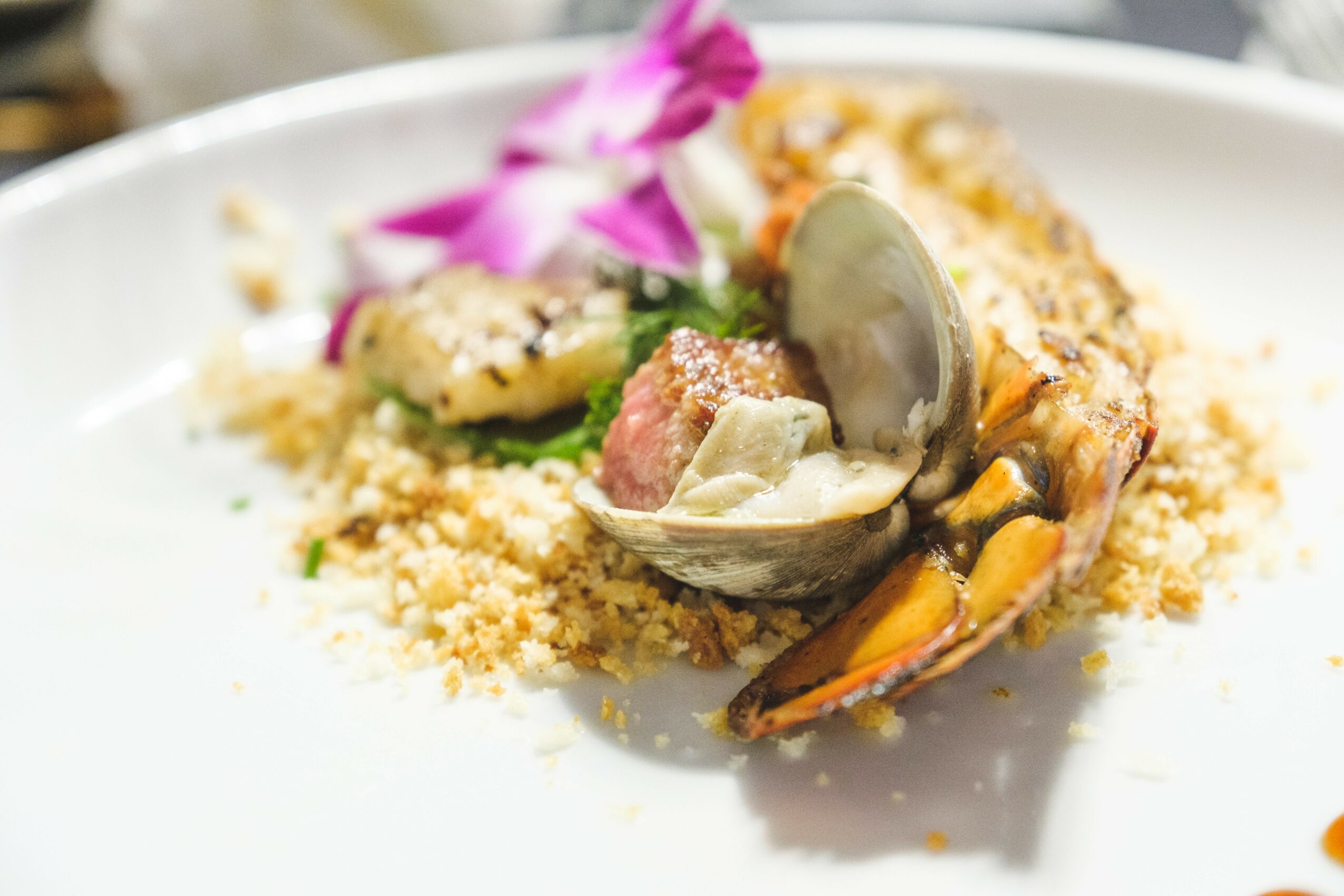 Seafood dish with mussels, shrimp, and garnishes on a white plate, with pink flower petals in the background.