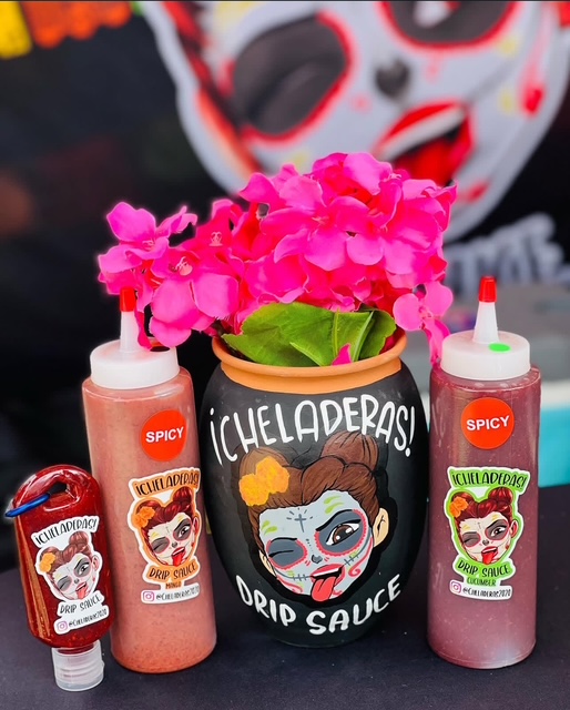 Pink flowers in a black pot with a cartoon face, surrounded by two bottles of spicy sauce, and a small container, all on a surface. A person with a painted face is in the background.