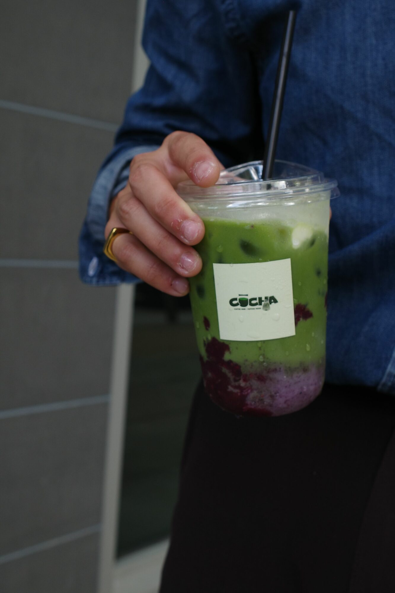 Person holding a green and purple smoothie in a clear cup with a straw, label on the cup, in front of a tiled wall.