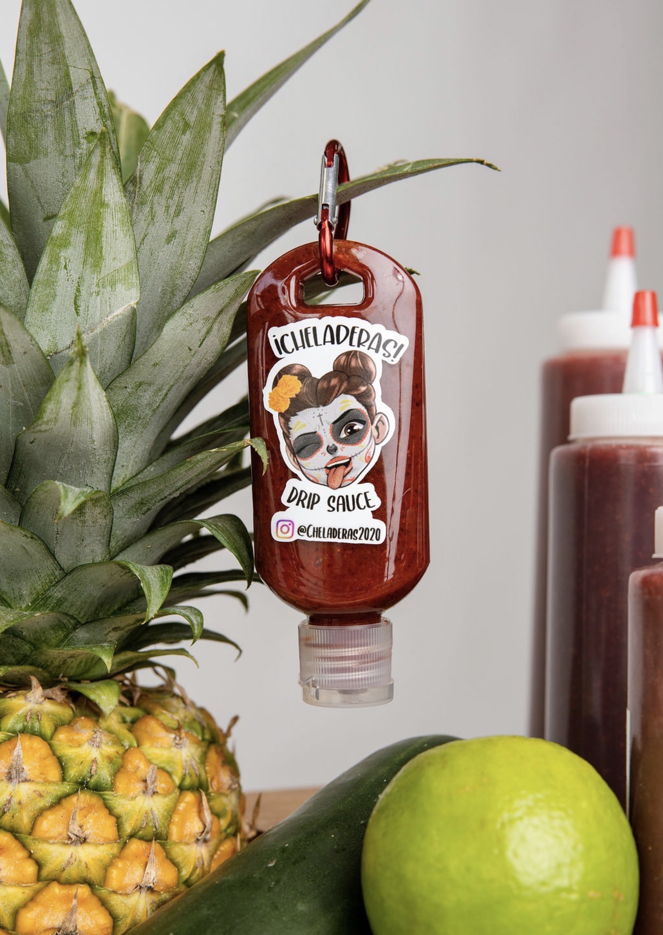 Bottle of drop sauce hanging from pineapple leaves, with lime and condiment bottles nearby, against a plain background.