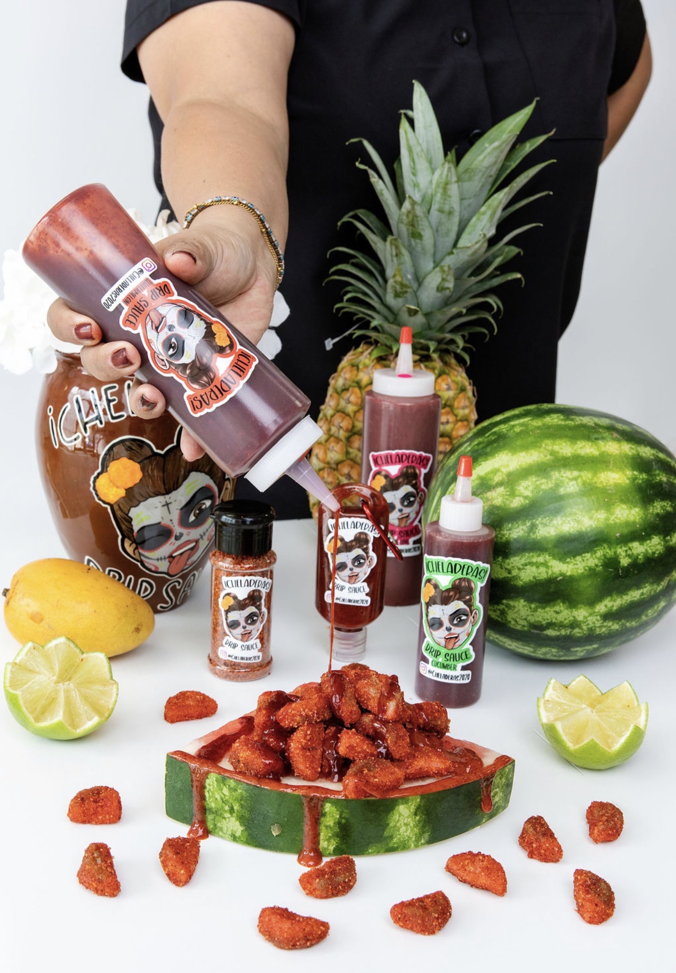 Person holding a bottle of sauce over a watermelon with sliced lemon, pineapple, and spicy food, surrounded by condiments and a pineapple.