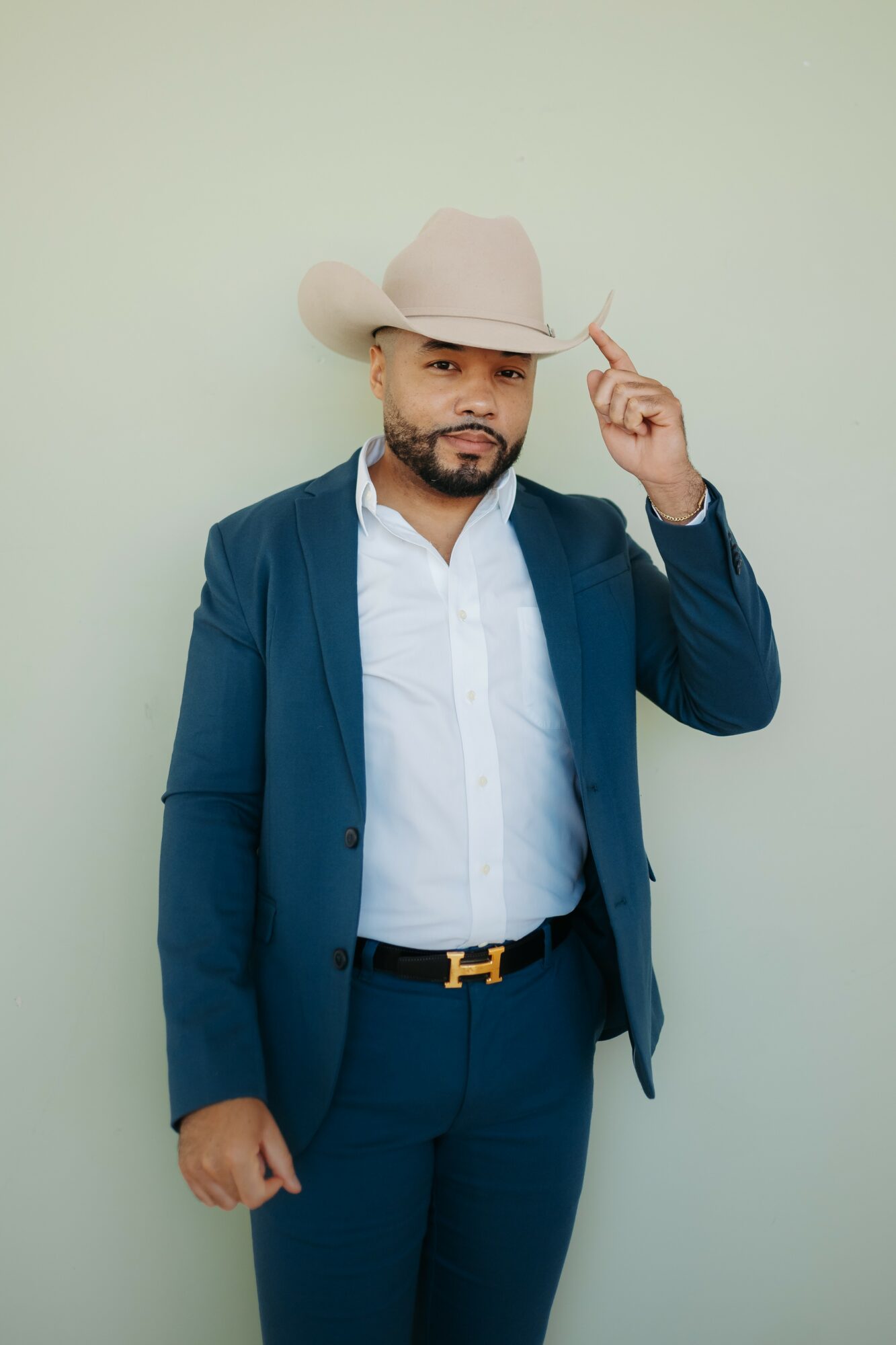 Man in a blue suit and white shirt wearing a large beige cowboy hat, pointing upward with his right hand.