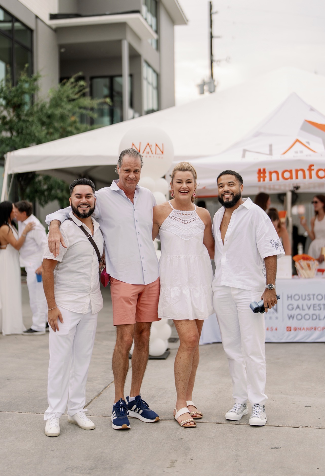 Four people stand together outdoors in front of a white tent, smiling, with a building and trees in the background.