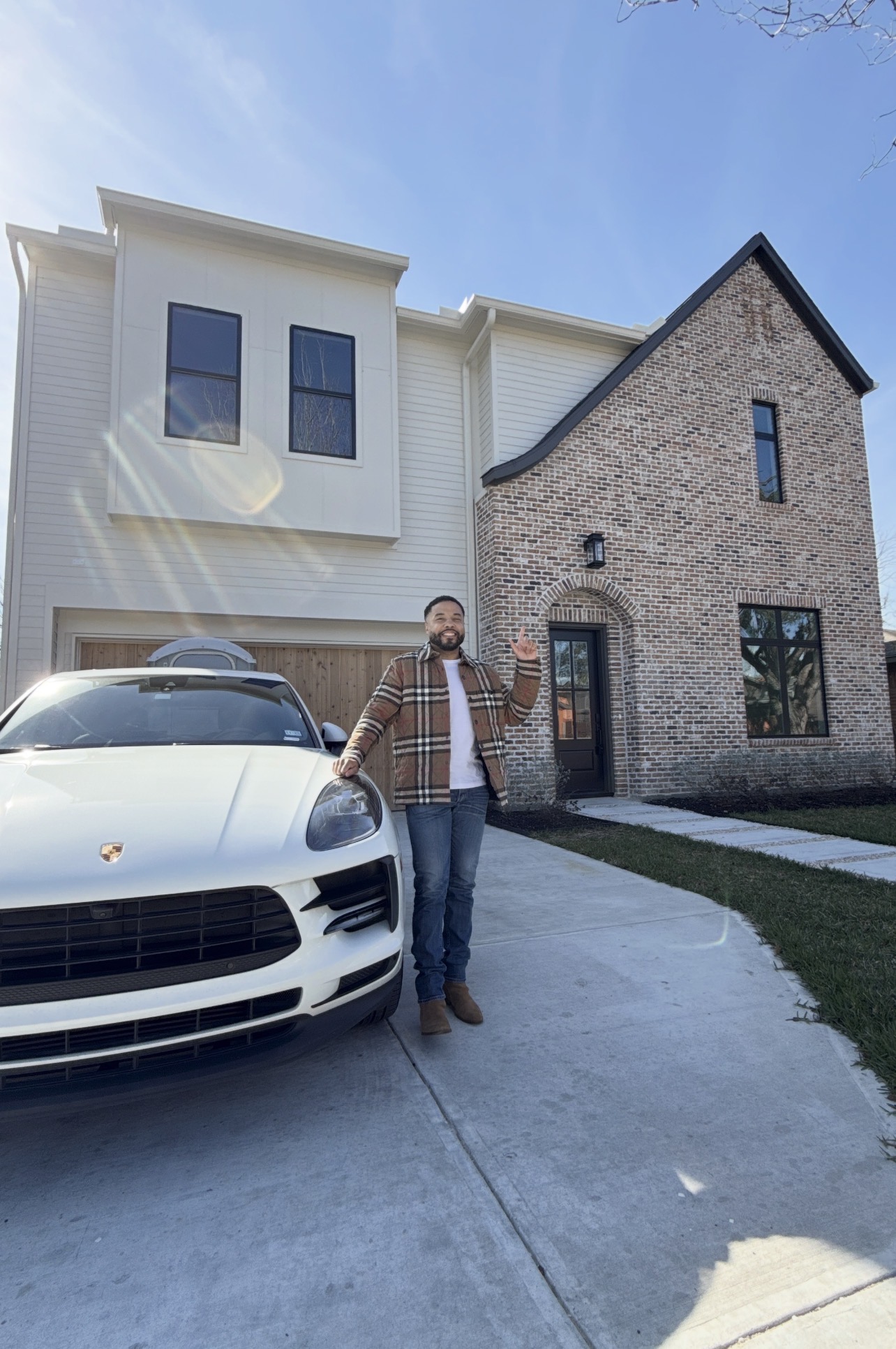 Person standing next to a white car in front of a modern house with brick and white siding, under a clear blue sky.