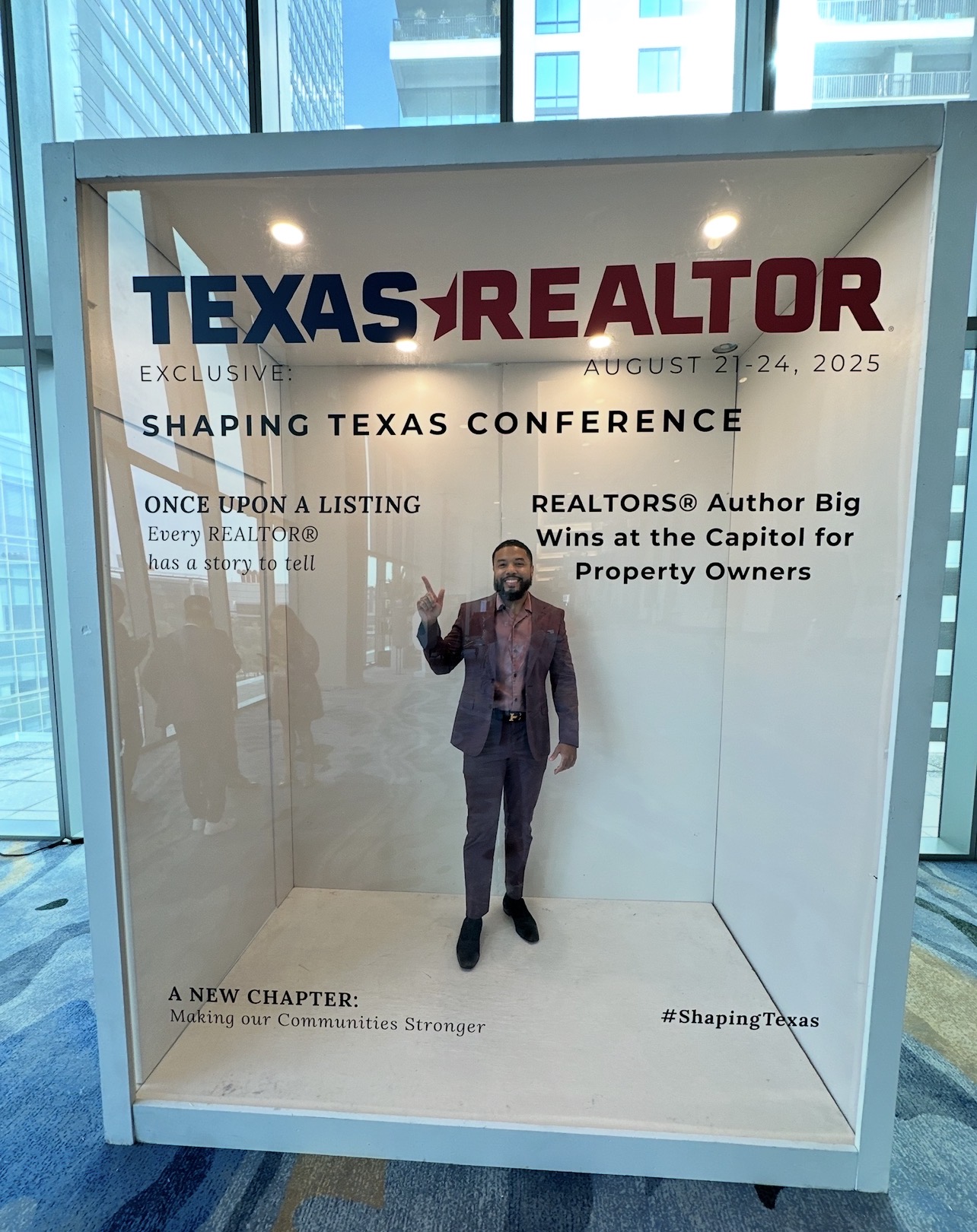 Person in suit standing inside a glass display case with a conference poster, in a modern building with large windows.