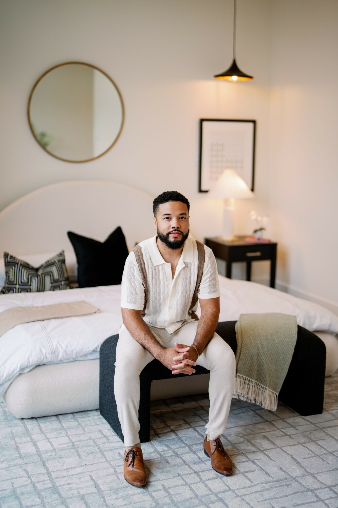 Man with beard and short hair sitting on a bed in a modern bedroom, with a round mirror and framed picture on the wall.