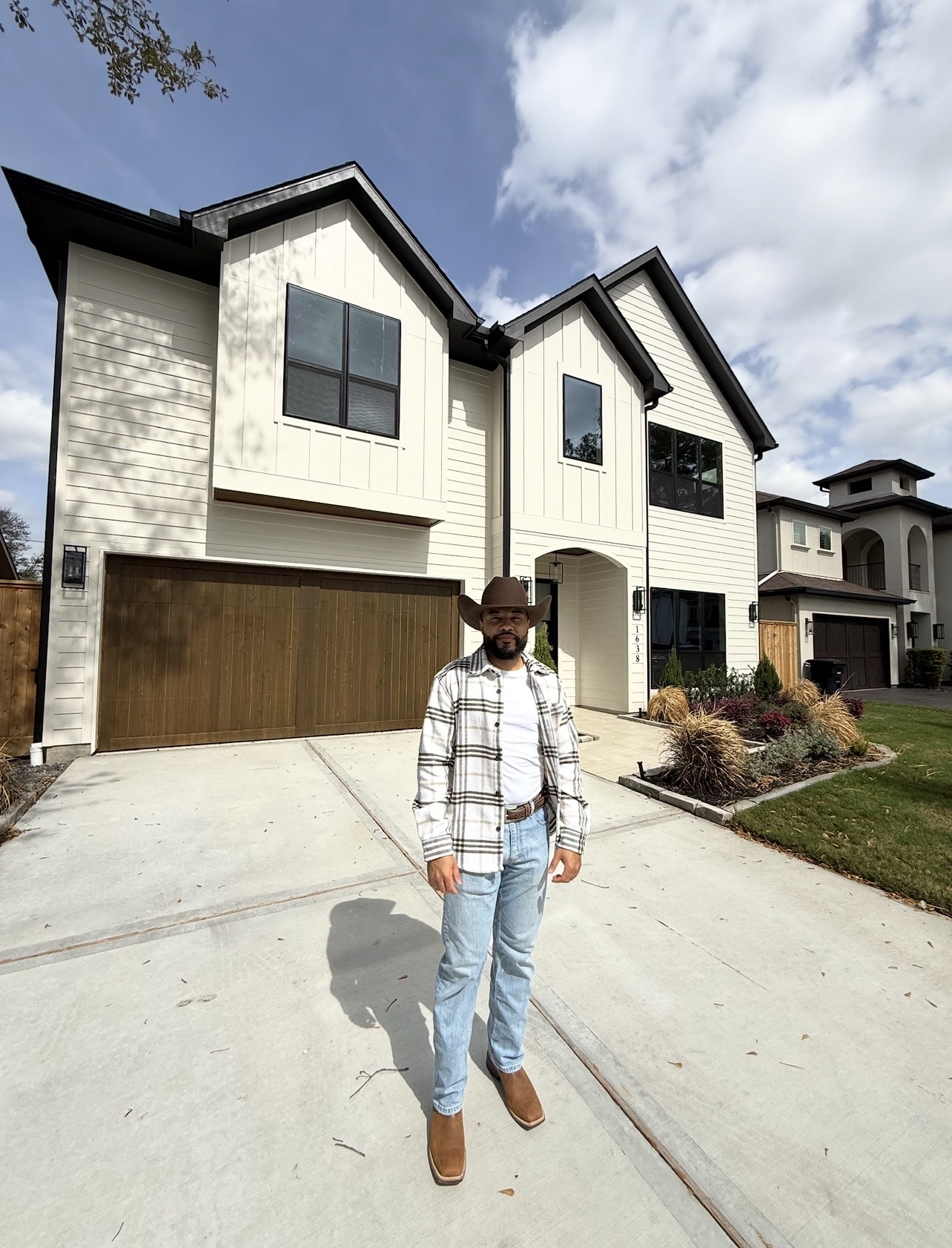 Person standing in front of a large, modern house with a garage, cloudy sky, and landscaped yard.